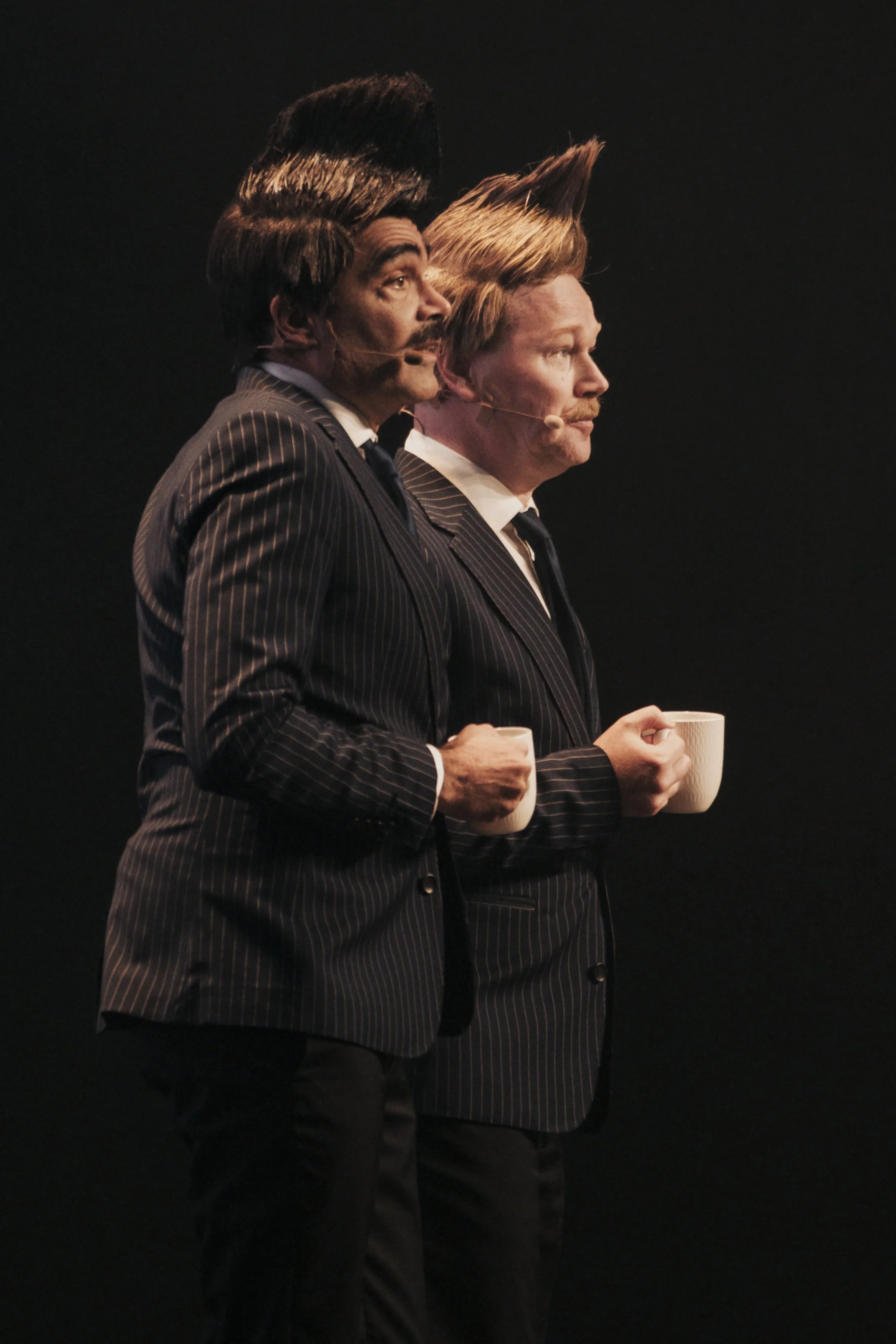 Två man i mörka kostymer med nummerlappar framför sig, håller koppar, står mot en svart bakgrund.
