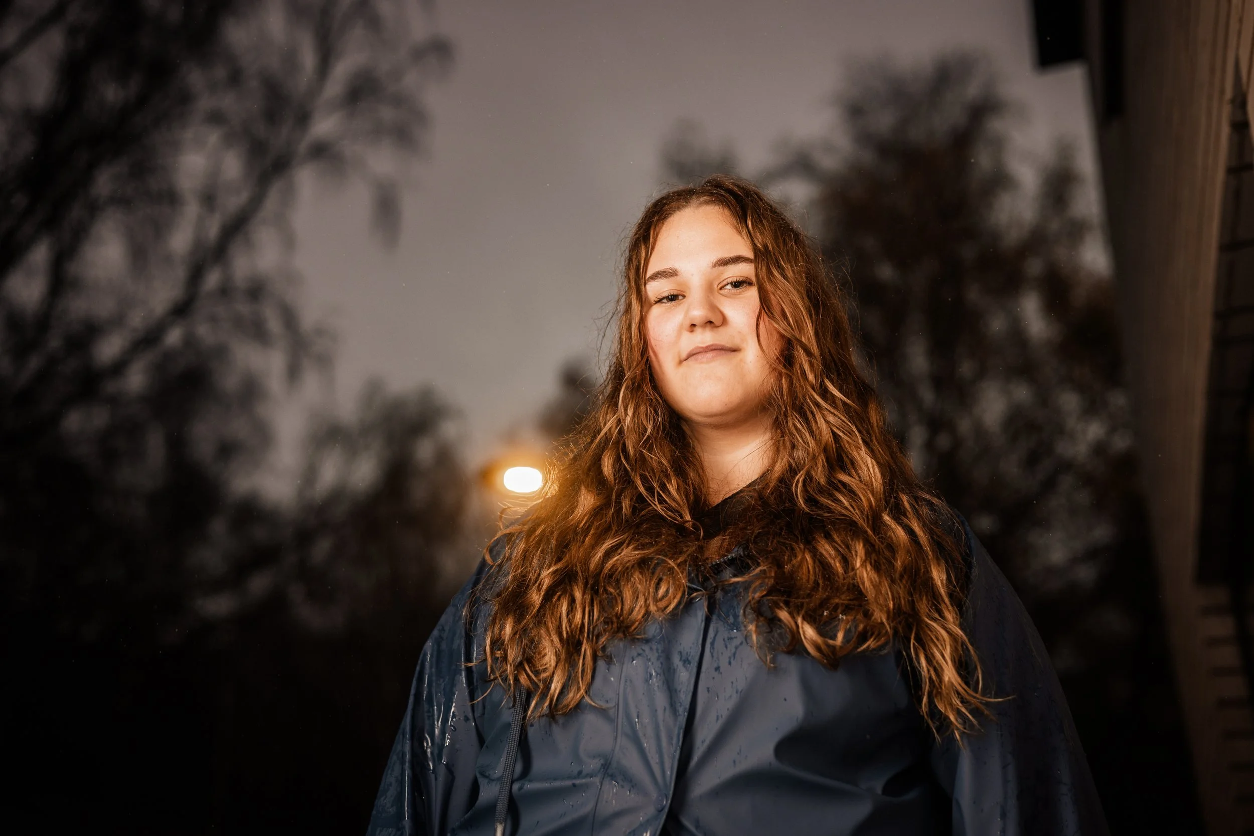 Ung kvinna med lockigt rödbrunt hår står utomhus på kvällen eller natten, med mörka träd och en gatlampa i bakgrunden.