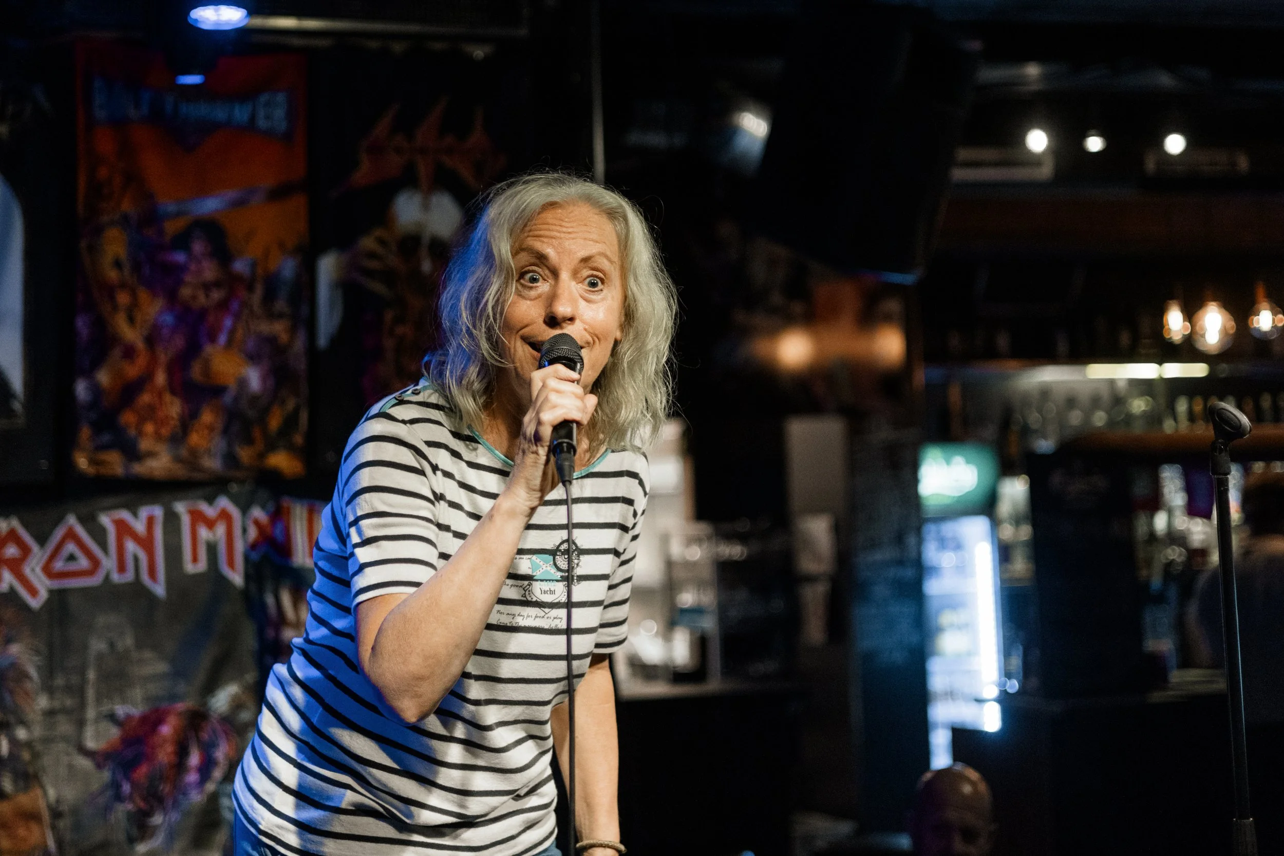 En äldre kvinna med blont, lockigt hår håller i en mikrofon och framträder i en bar eller klubb, med ett Iron Maiden-banner i bakgrunden.