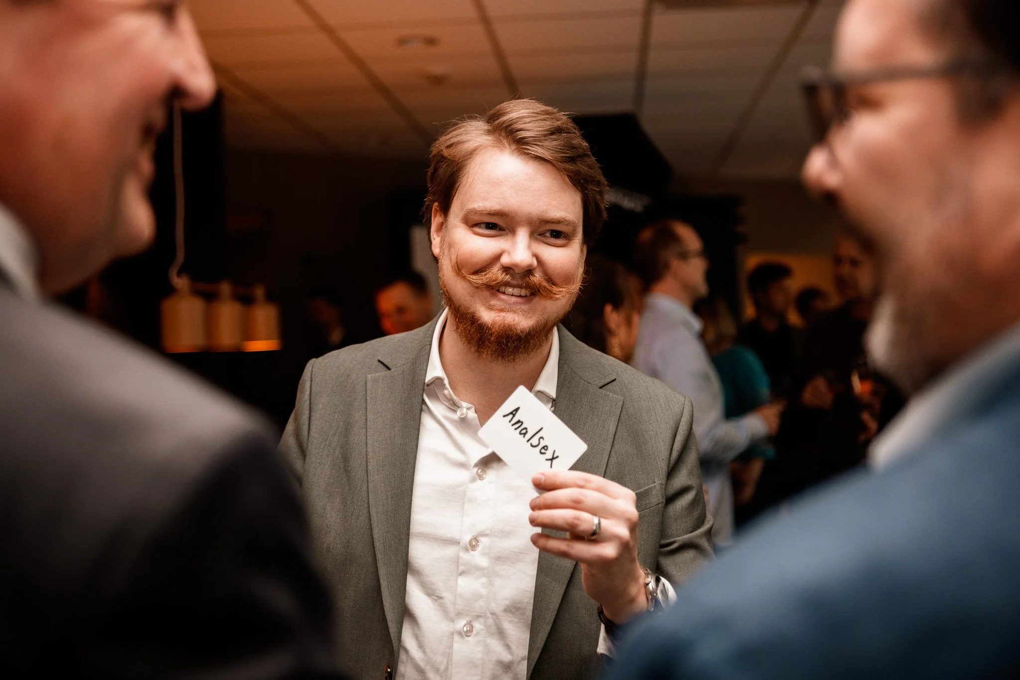 En man med lockigt hår och skägg ler och håller i ett namnplakat med texten 'Analsex'.