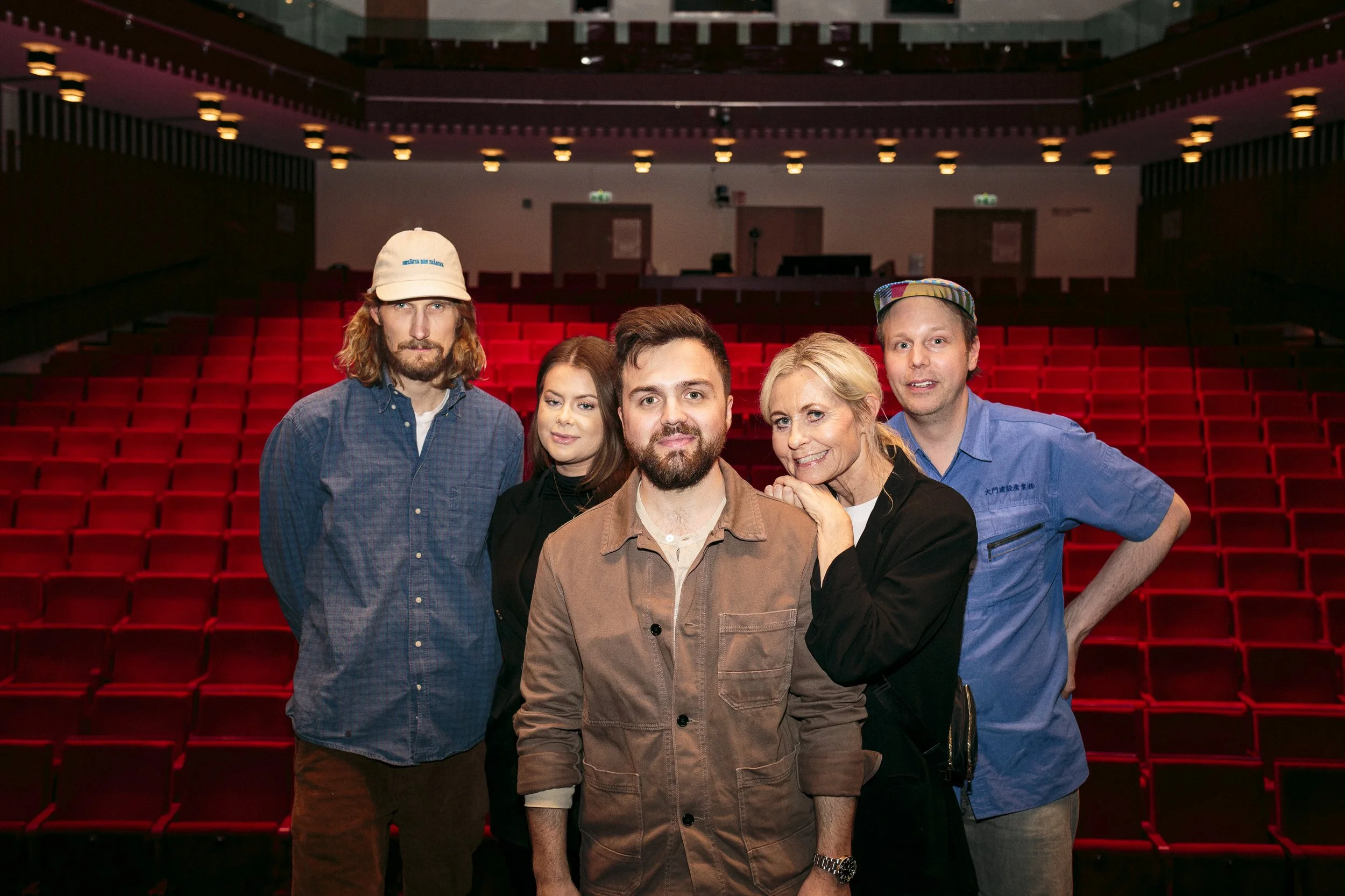 Fem personer i en tom teatersal med röda stolar, tre män och två kvinnor poserar för kameran.