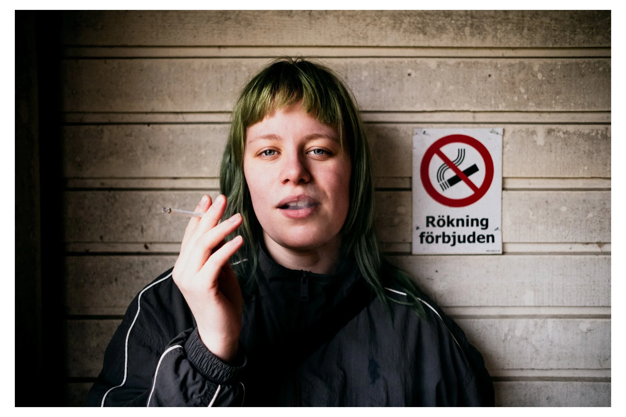 En ung person med grönt hår som håller en cigarett i handen står framför ett väggskydd med en förbudsskylt mot rökning.