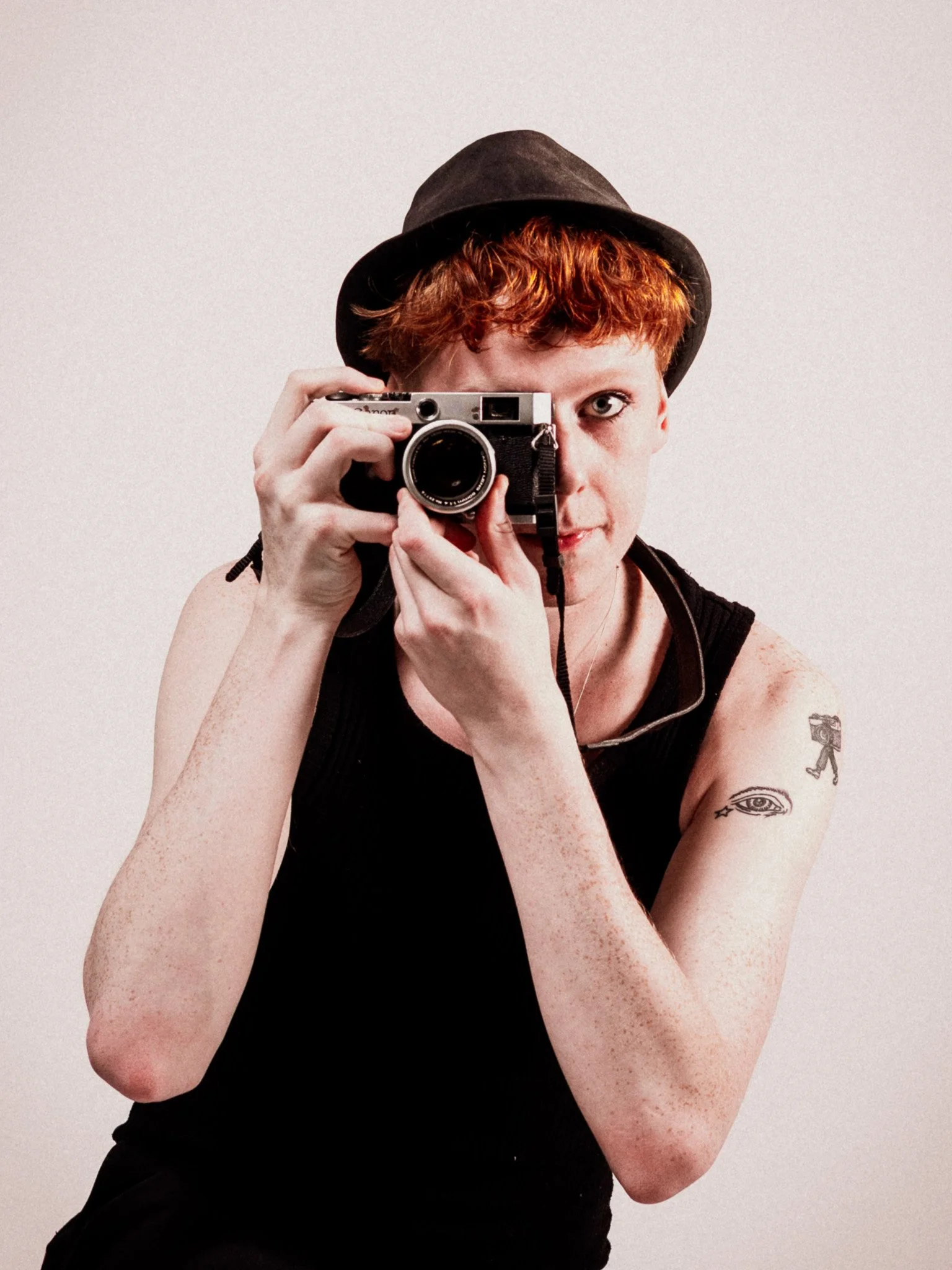 Person med rött hår, svart hatt, som tar ett självporträtt med en kamera, har tatueringar på armen, inklusive ett ögonmotiv.