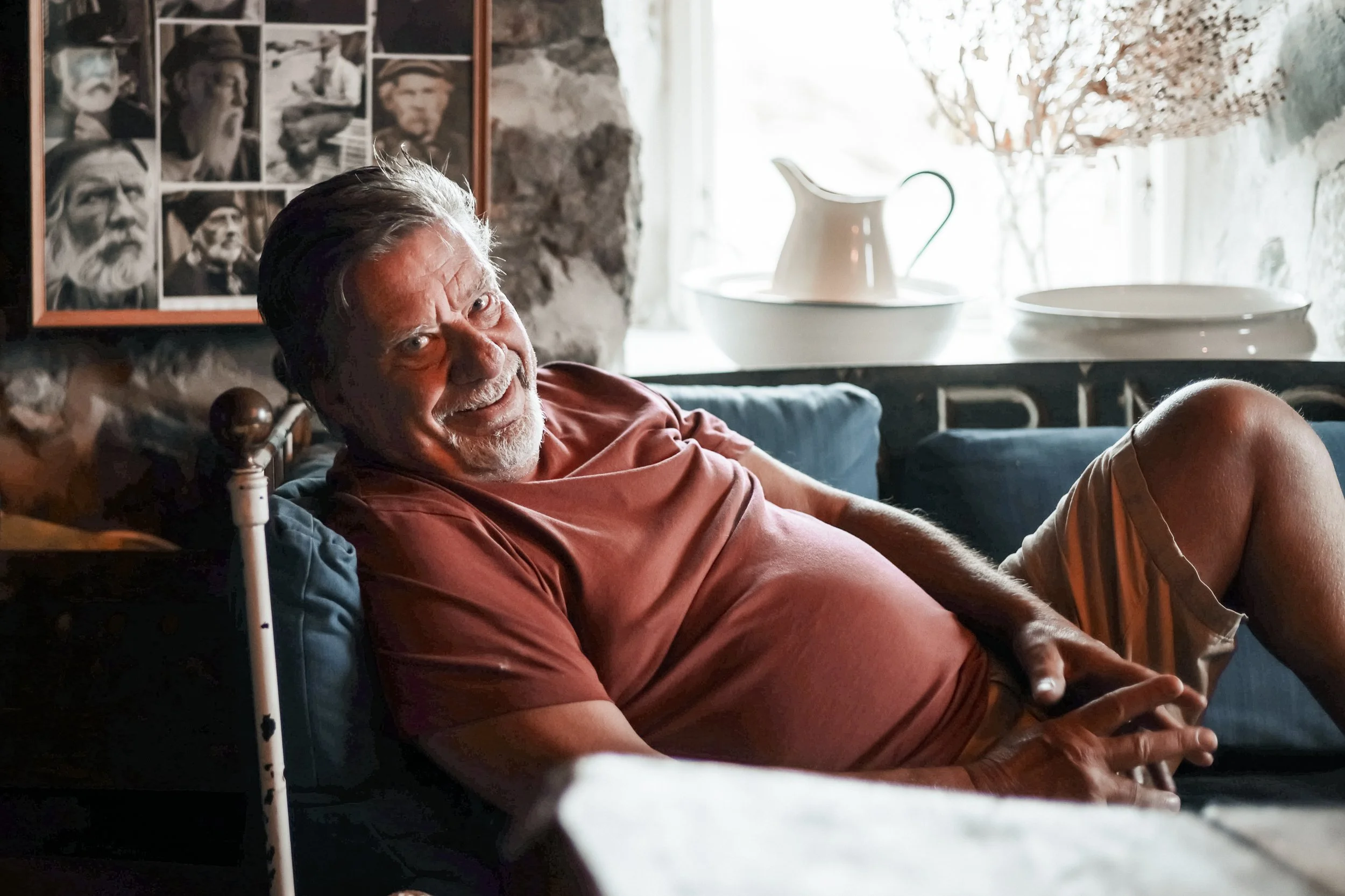 En äldre man med grått hår och skägg, ligger på en soffa och ler mot kameran. Han bär en röd t-shirt och beige shorts. Bakom honom syns ett fönster, några vita kärl och en växt med röda blad. På väggen till vänster finns en fotovägg med svartvita por