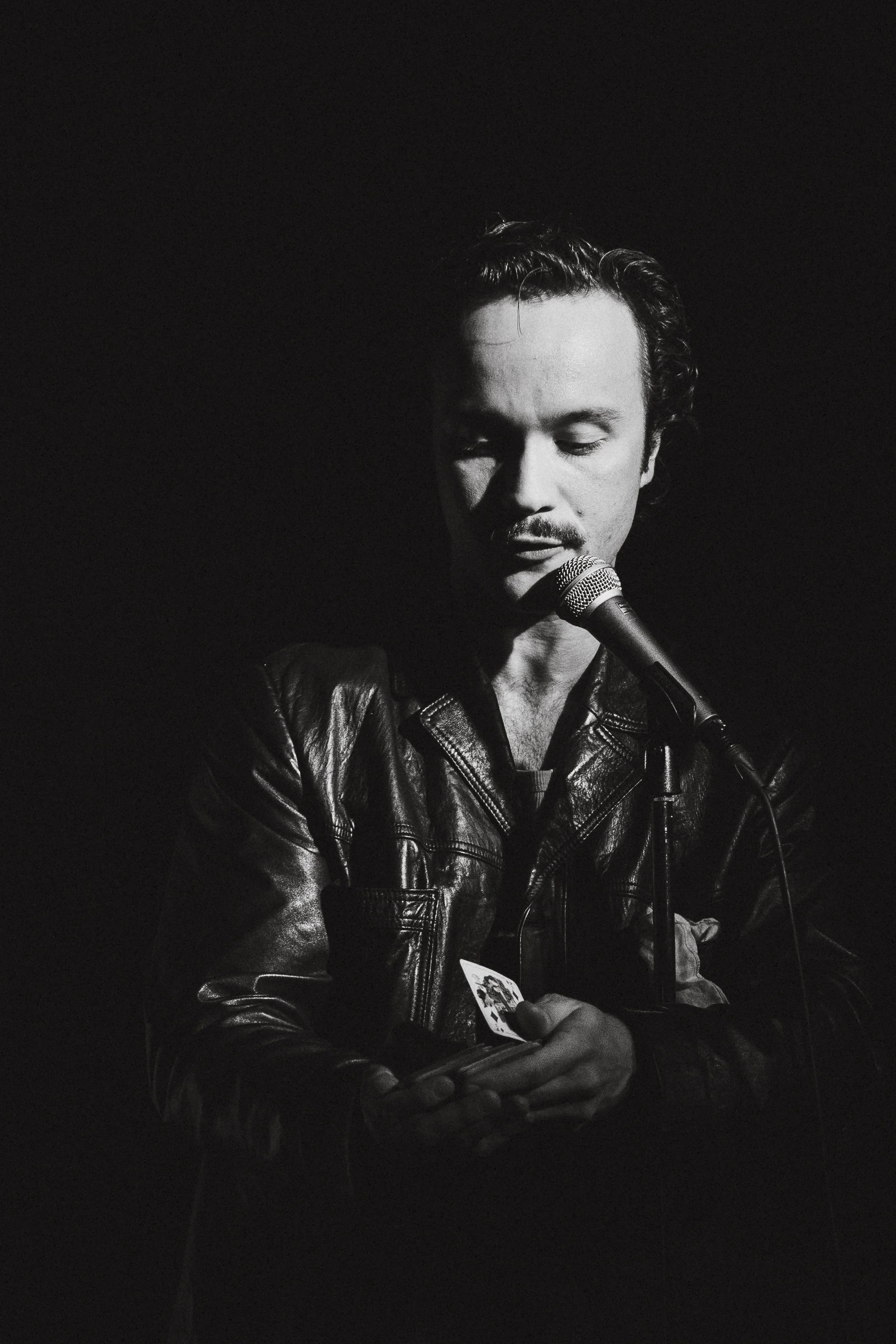 En man i läderjacka står framför en mikrofon, hans ögon är slut och han håller i ett kort eller ett förmodligen telefon.