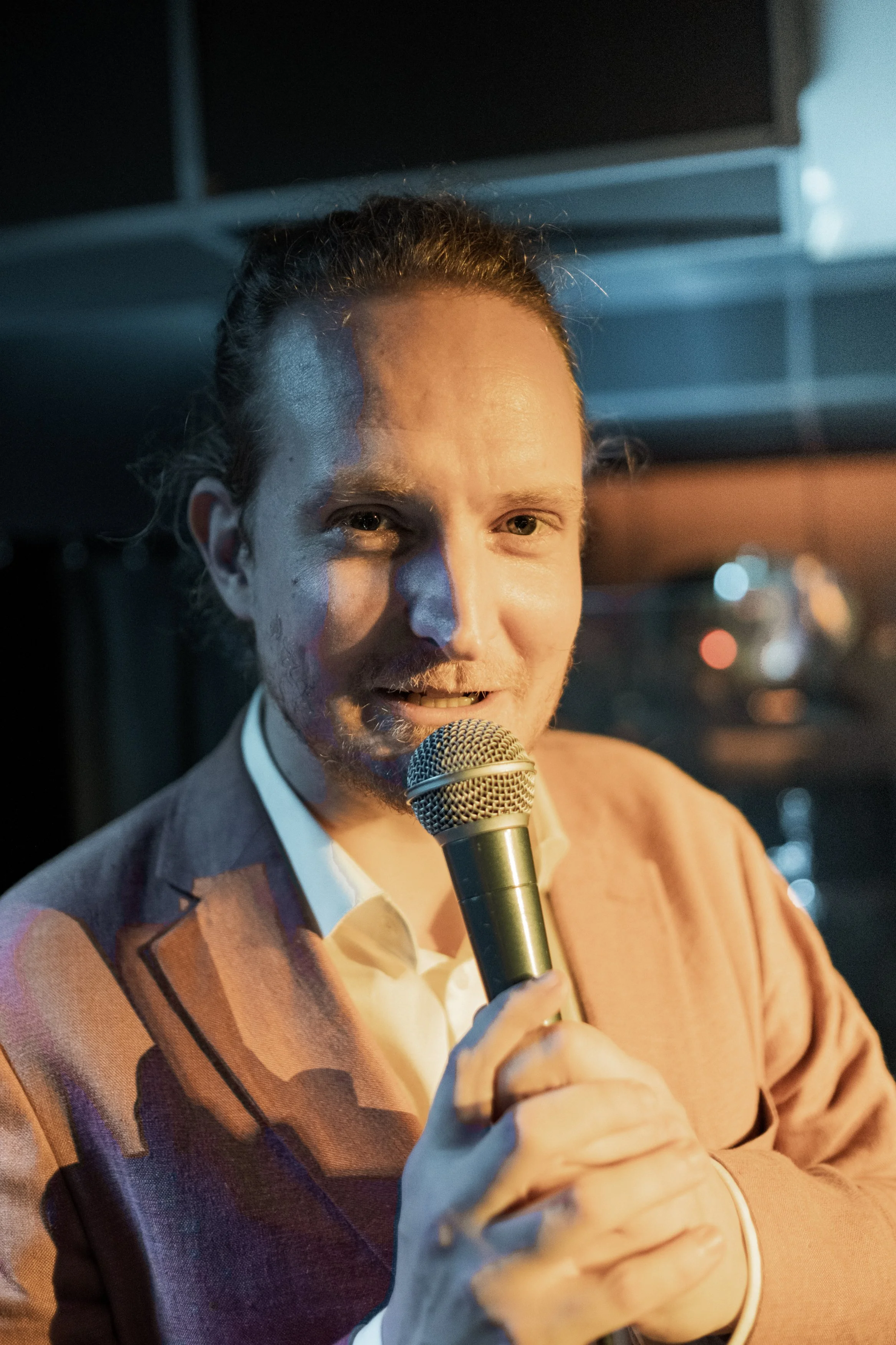 Man med långt hår i en hästsvans, klädd i en ljusrosa kavaj, talar i mikrofon under en kvällsmöte eller föreställning.