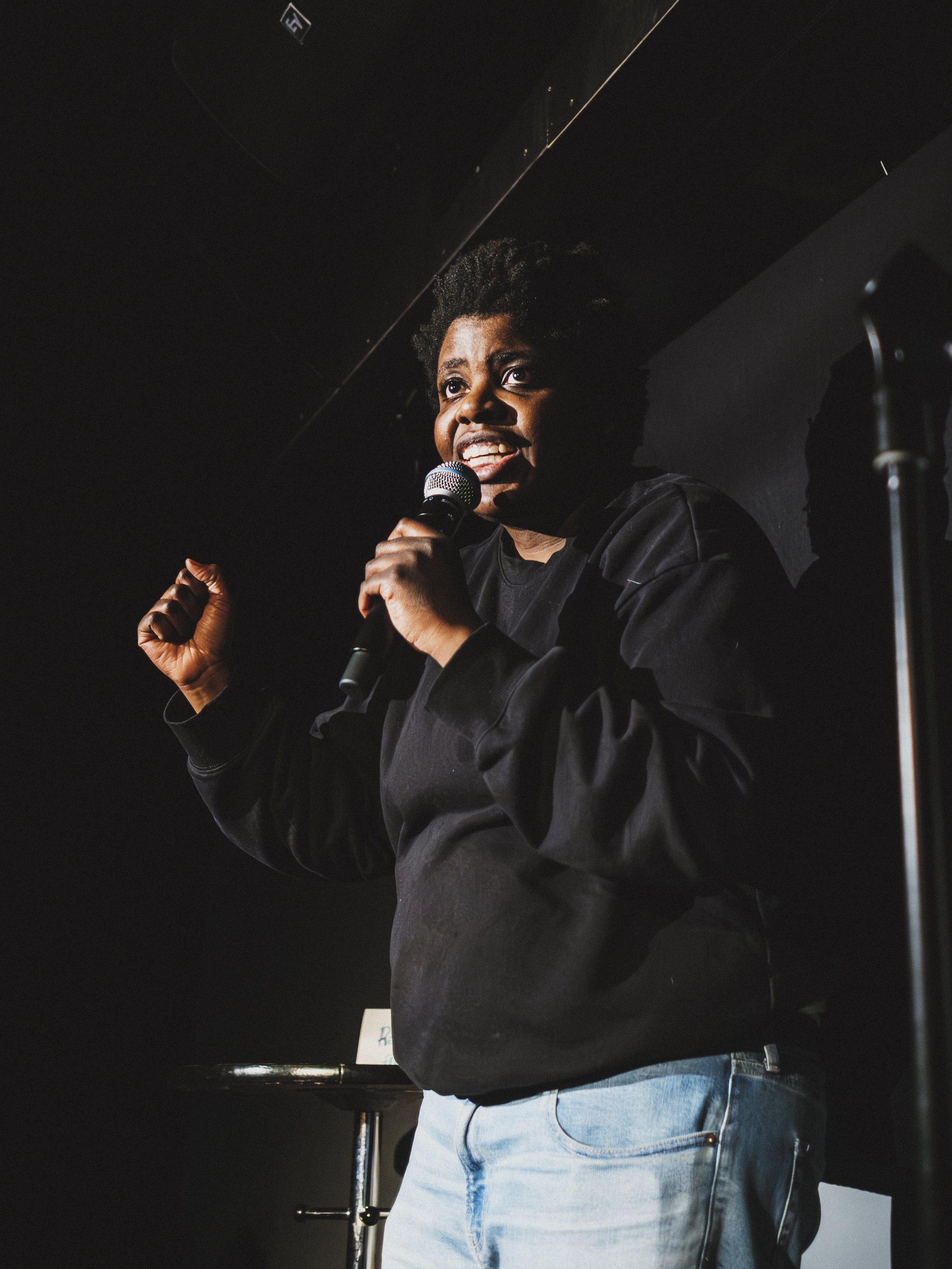 En kvinna håller en mikrofon och talar framför en mörk bakgrund, ser engagerad och entusiastisk ut.