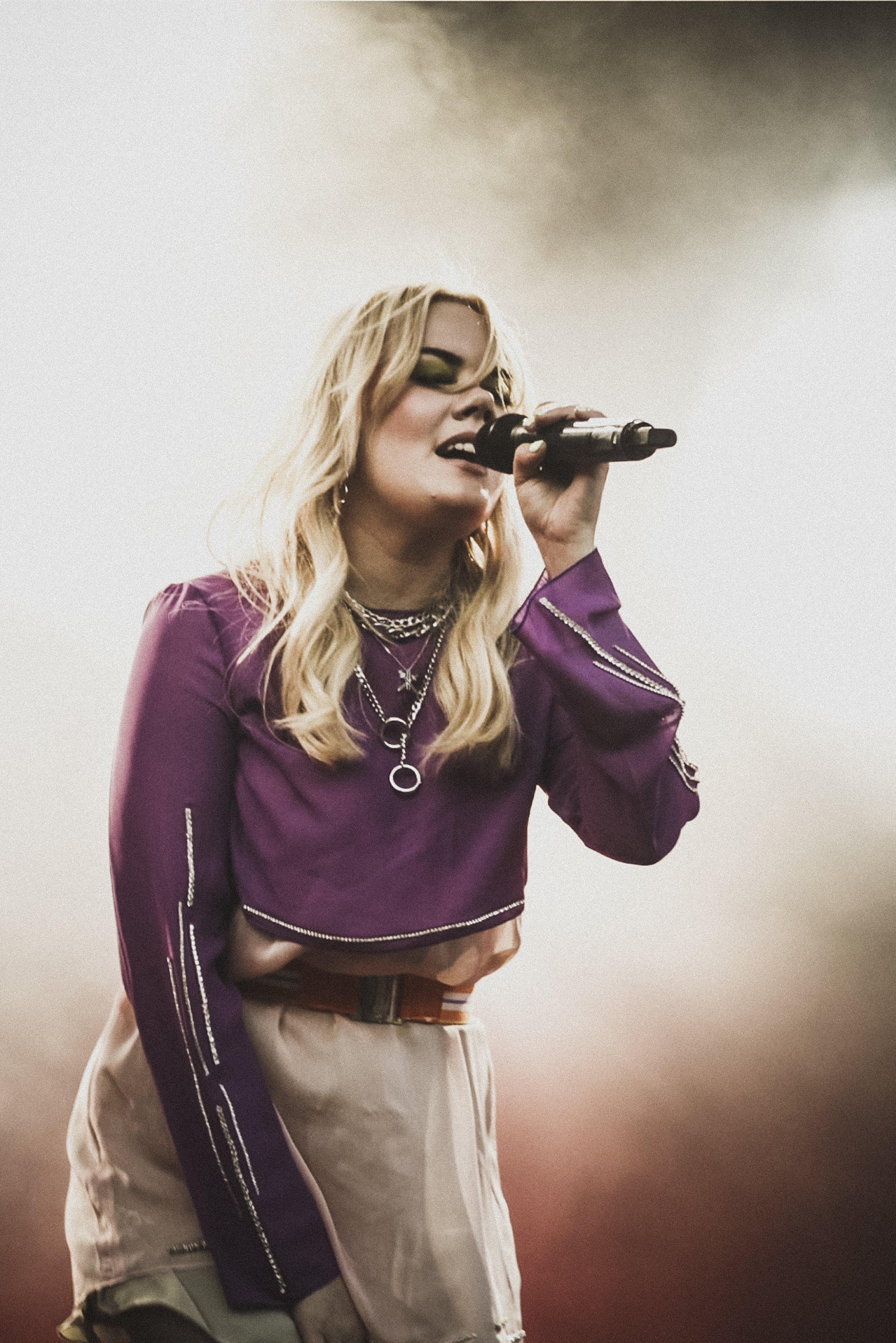 En ung kvinna med långt blont hår sjunger i mikrofon, bär en lila crop top med metalldetaljer och ljusfärgade byxor, omgiven av dimmigt ljus.