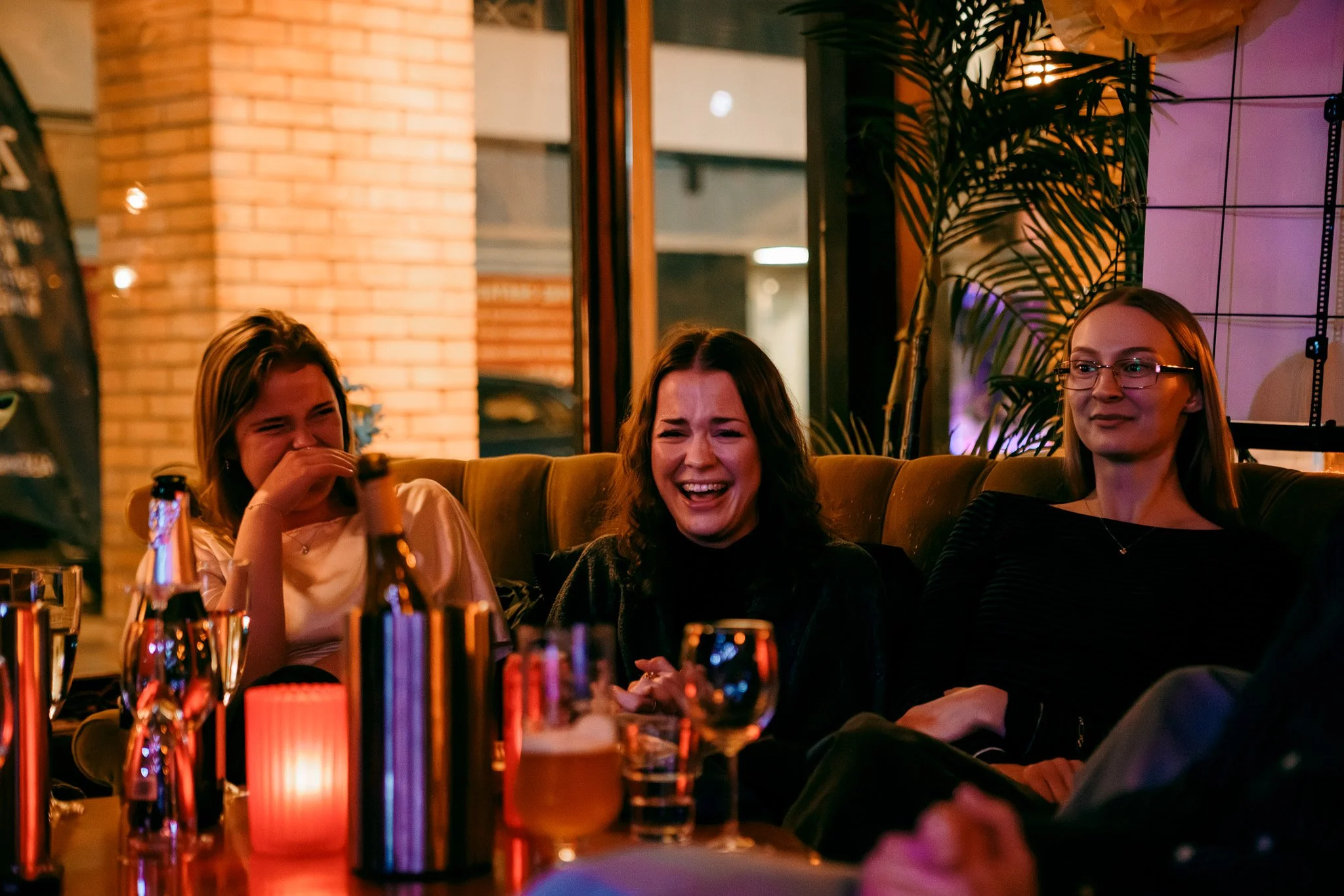 Tre kvinnor skrattar och har trevligt i en bar eller restaurang med varma belysningar och växtdekorationer.