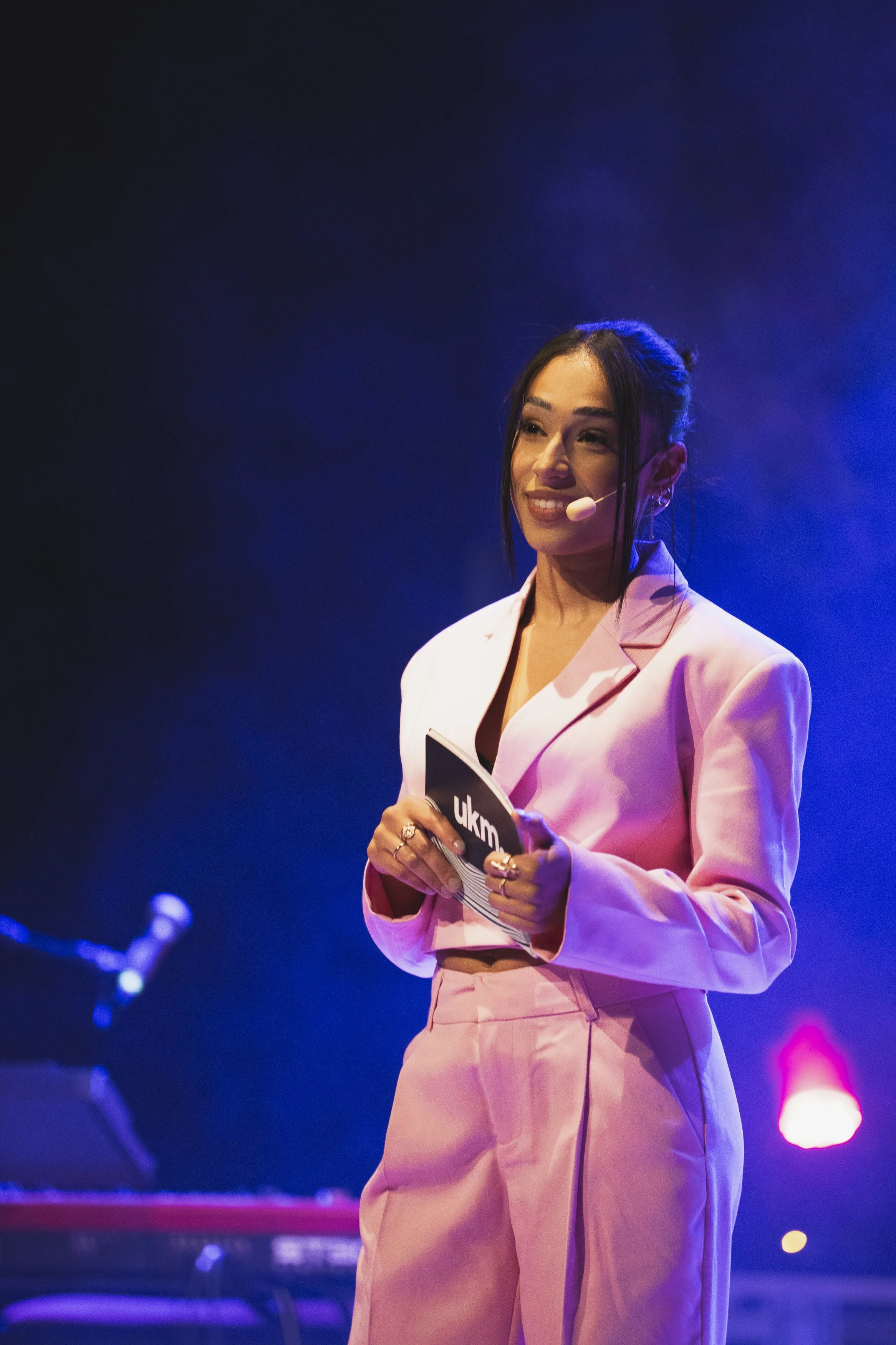 En kvinna i rosa kostym står på scen med mikrofon och papper, bakom henne syns en keyboard och mörk bakgrund med blå och rosa ljus.