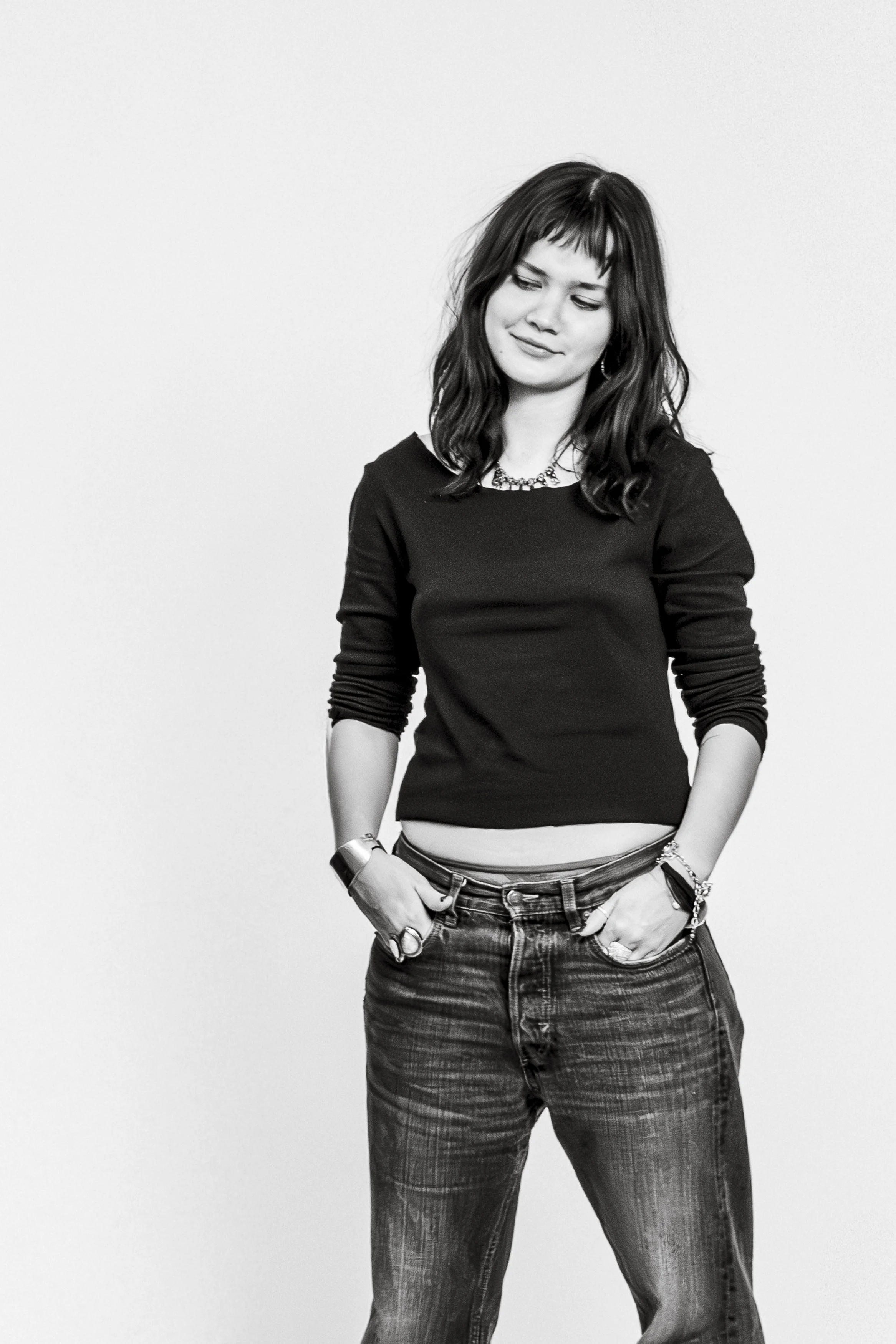 Svartvit bild av en ung kvinna med mörkt, axellångt hår och lugg, som står med händerna i fickorna och tittar lätt nedåt, klädd i svart tröja och jeans.