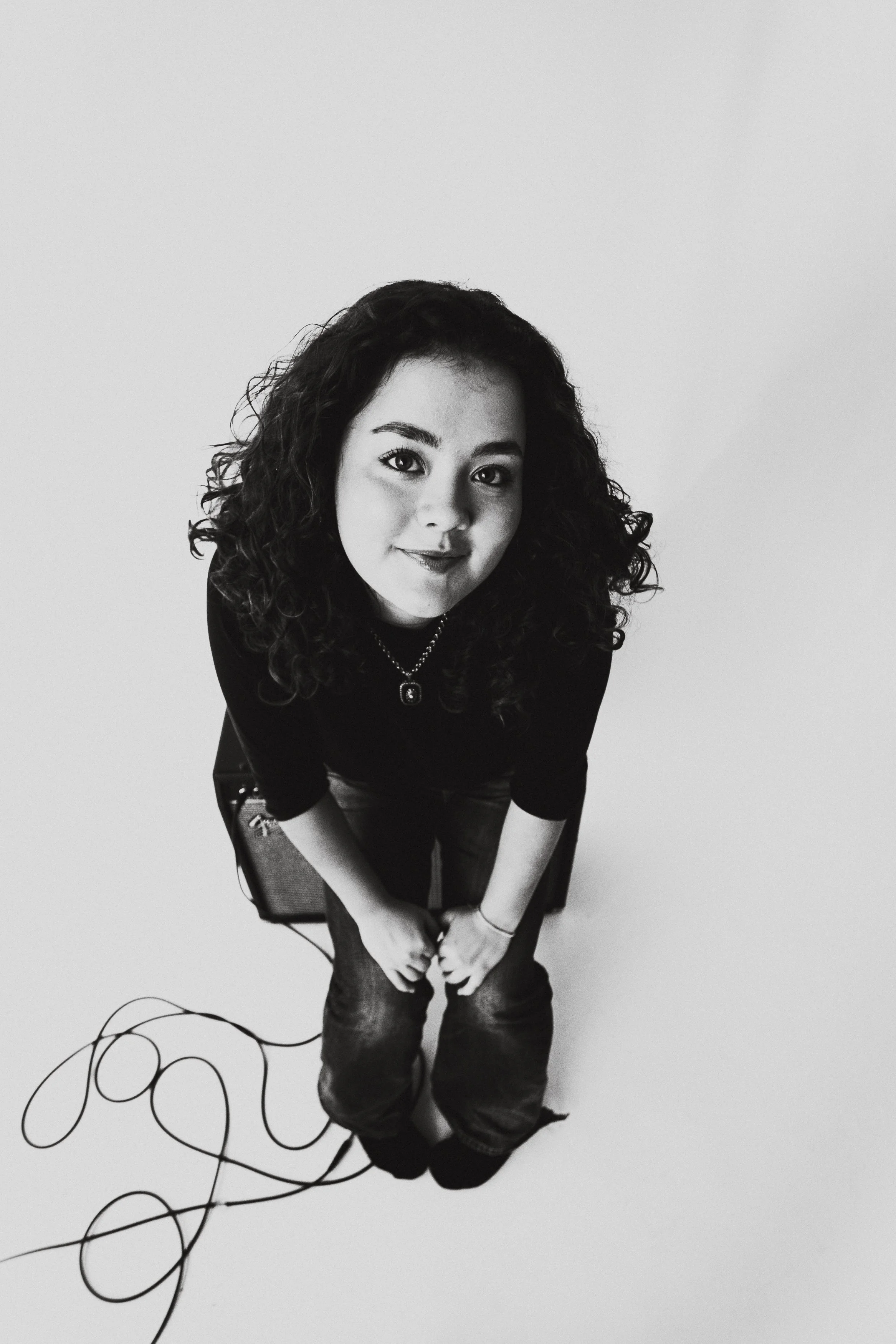 En svartvit bild av en kvinna med lockigt hår som sitter på en stol. Hon tittar upp mot kameran och bär ett halsband och en svart tröja.