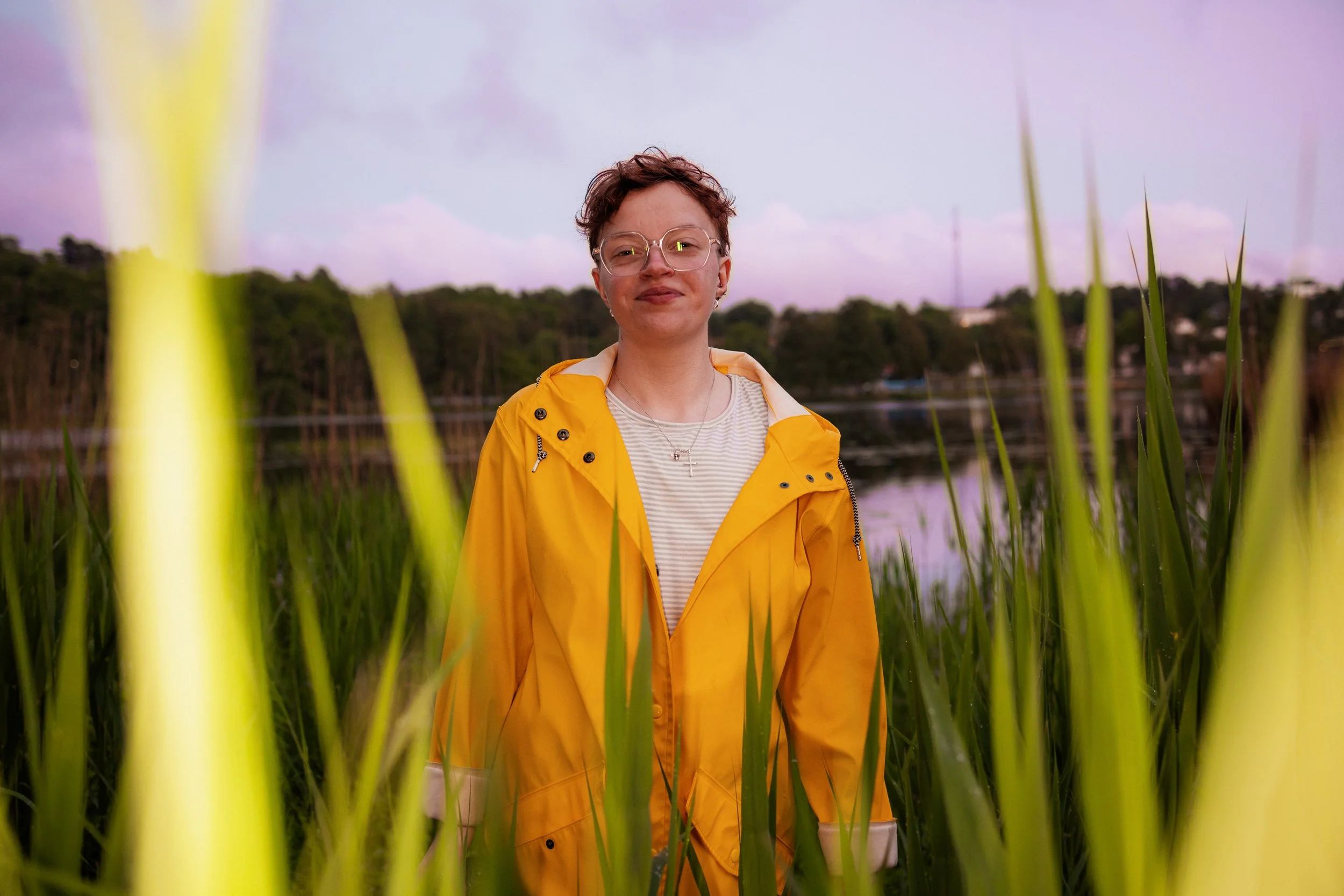 En person i gul regnjacka står bland grönska med vatten och skog i bakgrunden, under en kvällshimmel