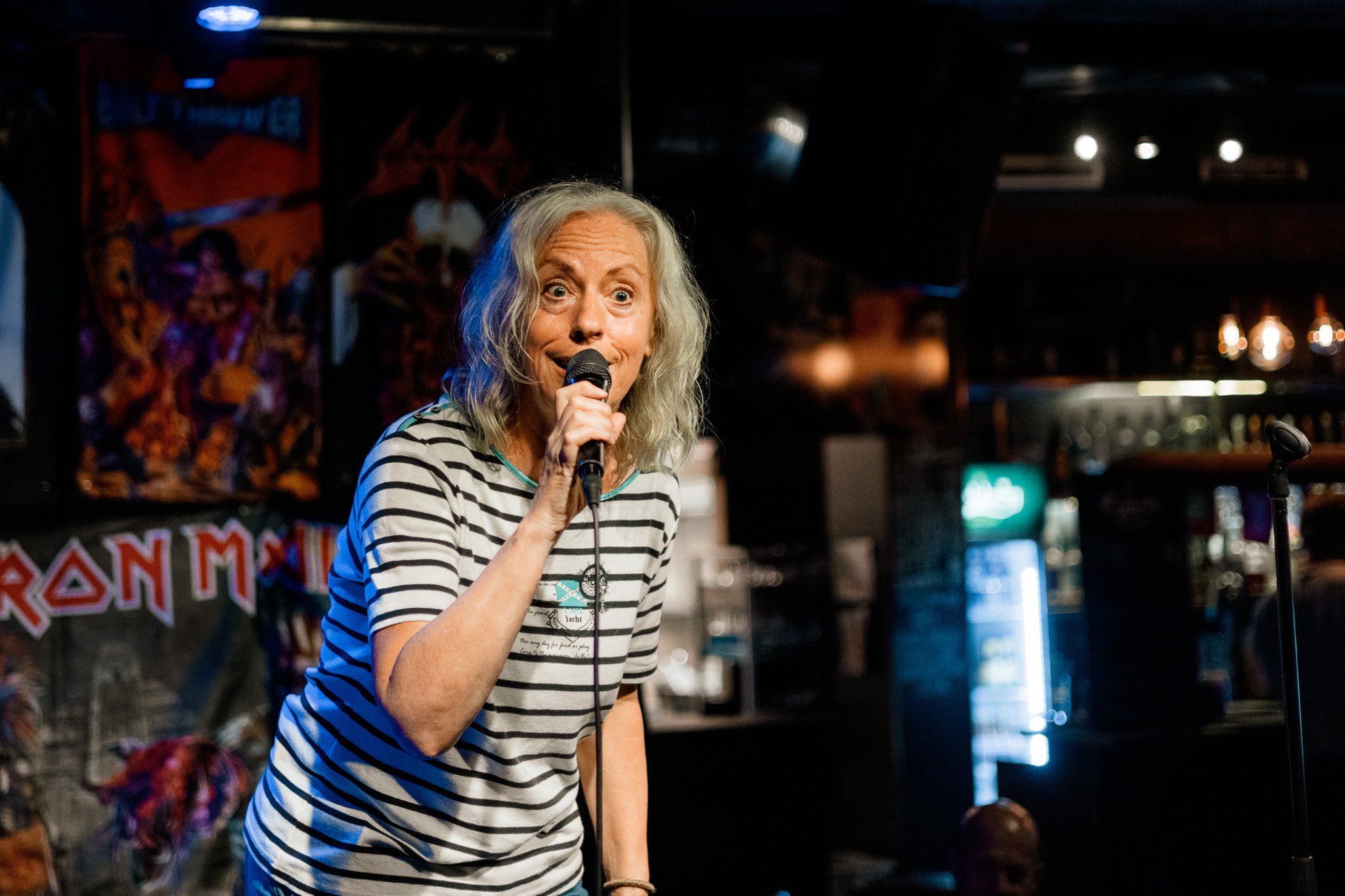 En kvinna med blont, vågigt hår håller i en mikrofon och tittar mot kameran, hon står i ett mörkt rum med discobollar och en Bar i bakgrunden.