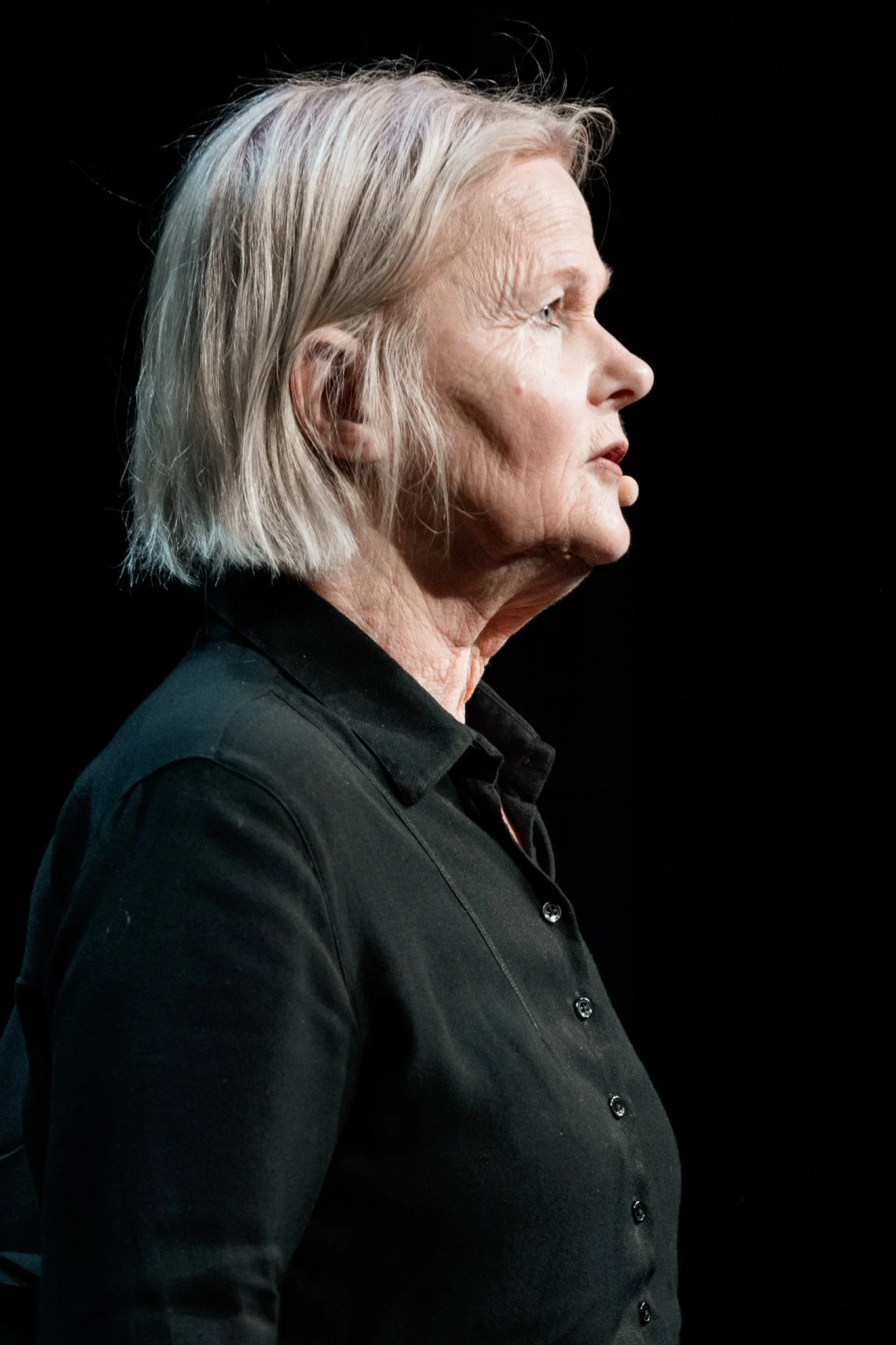 Profilbild av en äldre kvinna med grått hår, iförd svart skjorta, mot en svart bakgrund.