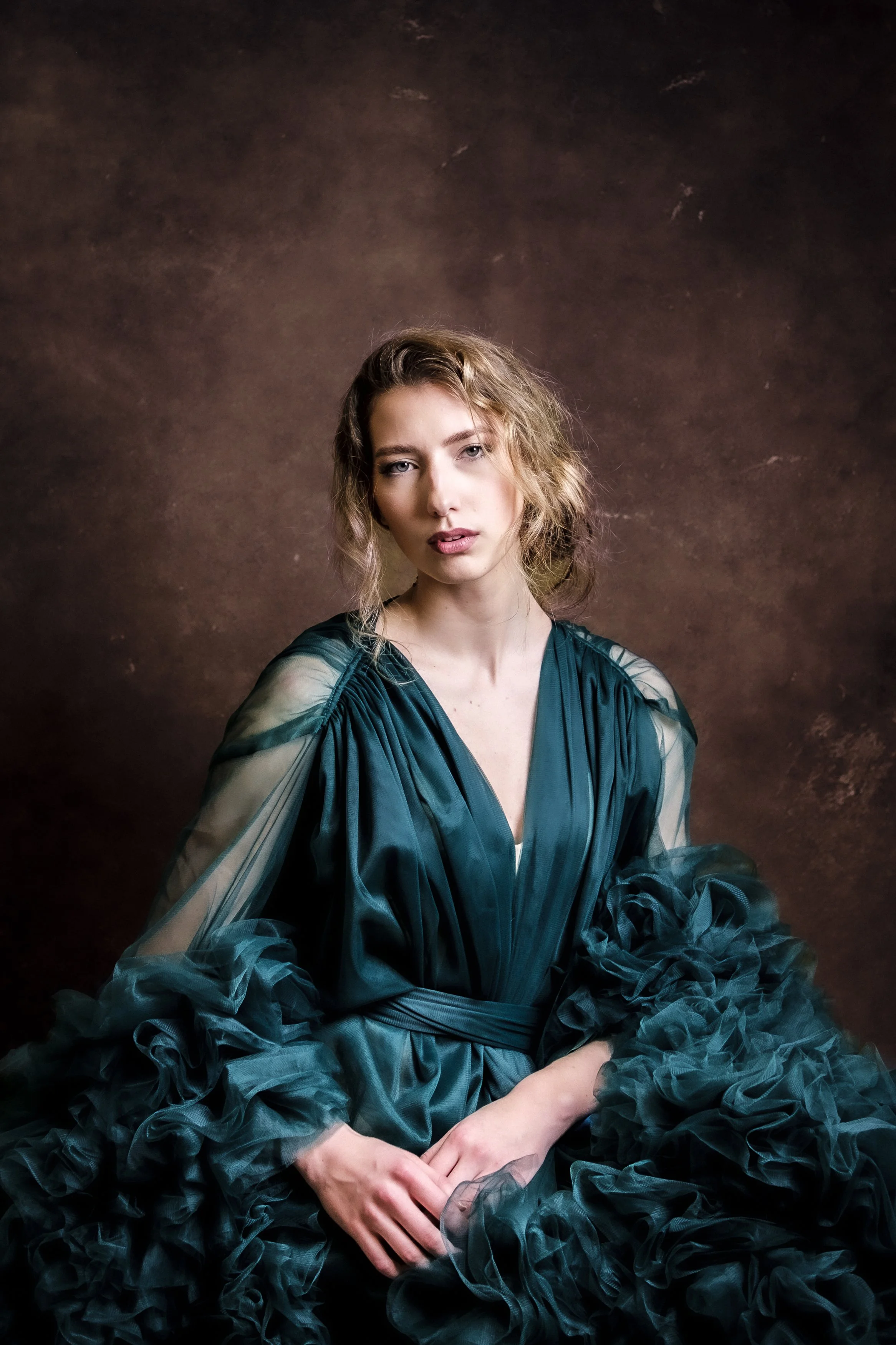 Porträtt av en ung kvinna med lockigt hår i en elegant mörkblå klänning med voluminösa ärmar, sittande mot en brun bakgrund.