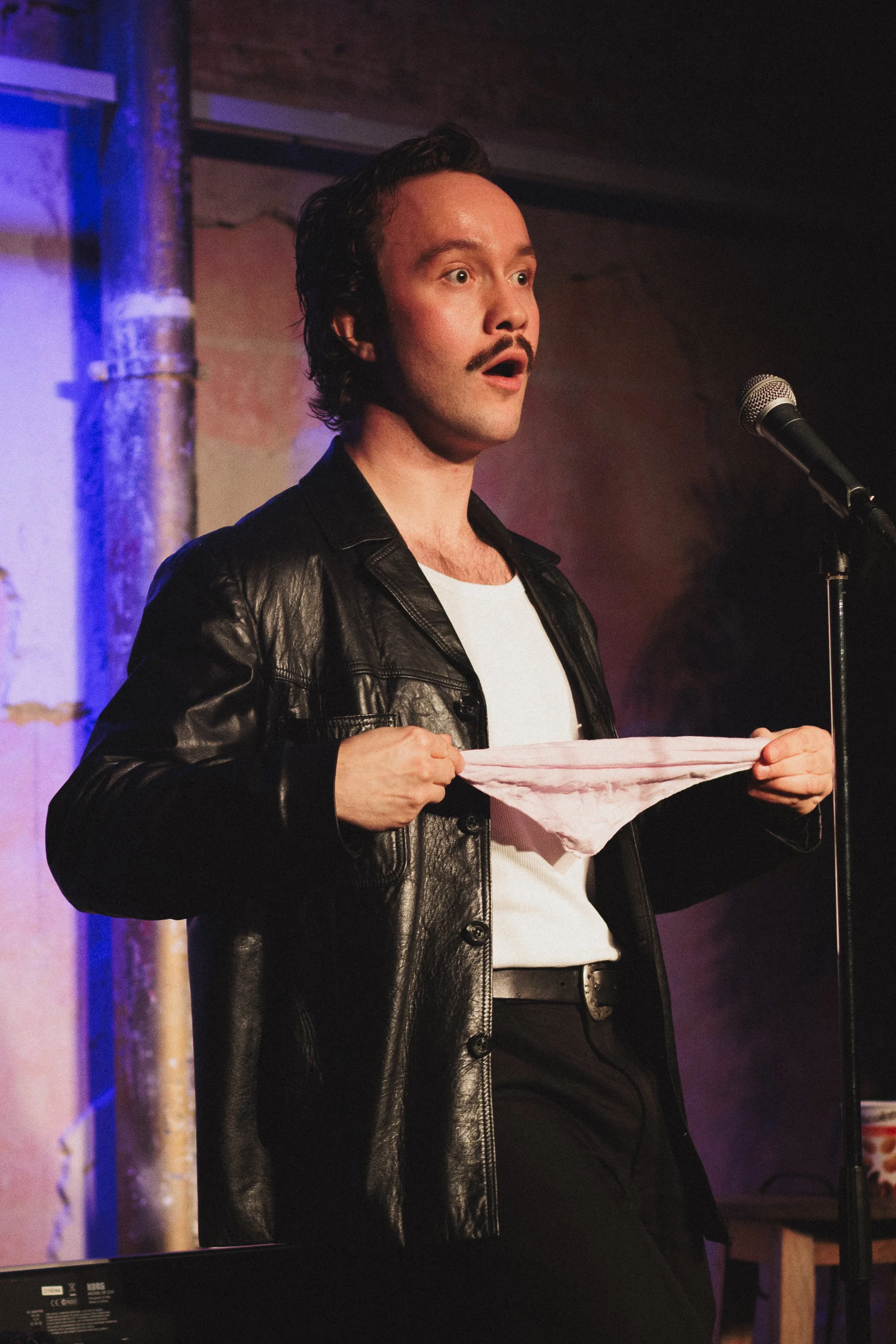 Man på scen med mustasch, svart läderjacka, vita T-shirt, håller i rosa underkläder, mimar förvånad.