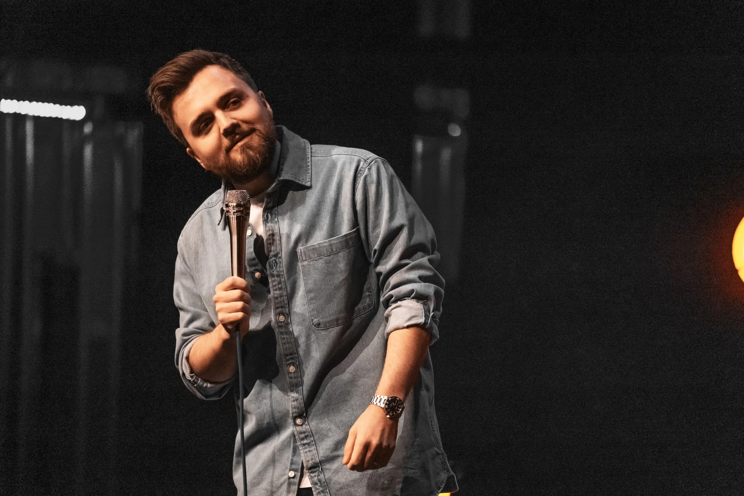 En man håller en mikrofon på scenen, klädd i en blå jeansjacka och har ett leende.