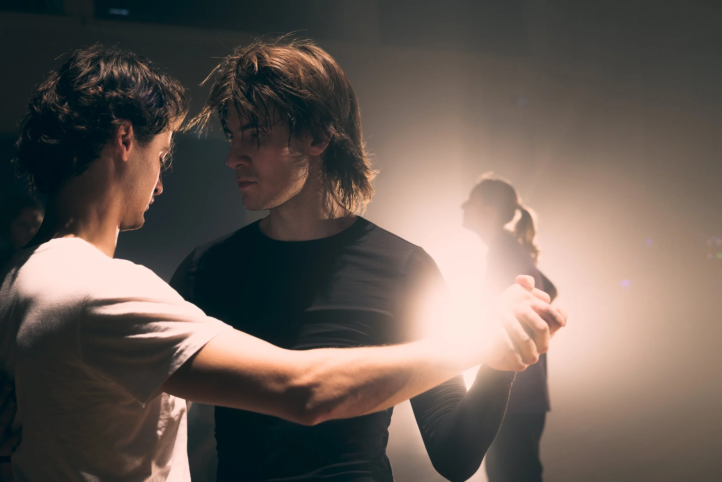 Två unga män dansar nära varandra i ett mörkt rum med ett starkt ljus bakifrån.