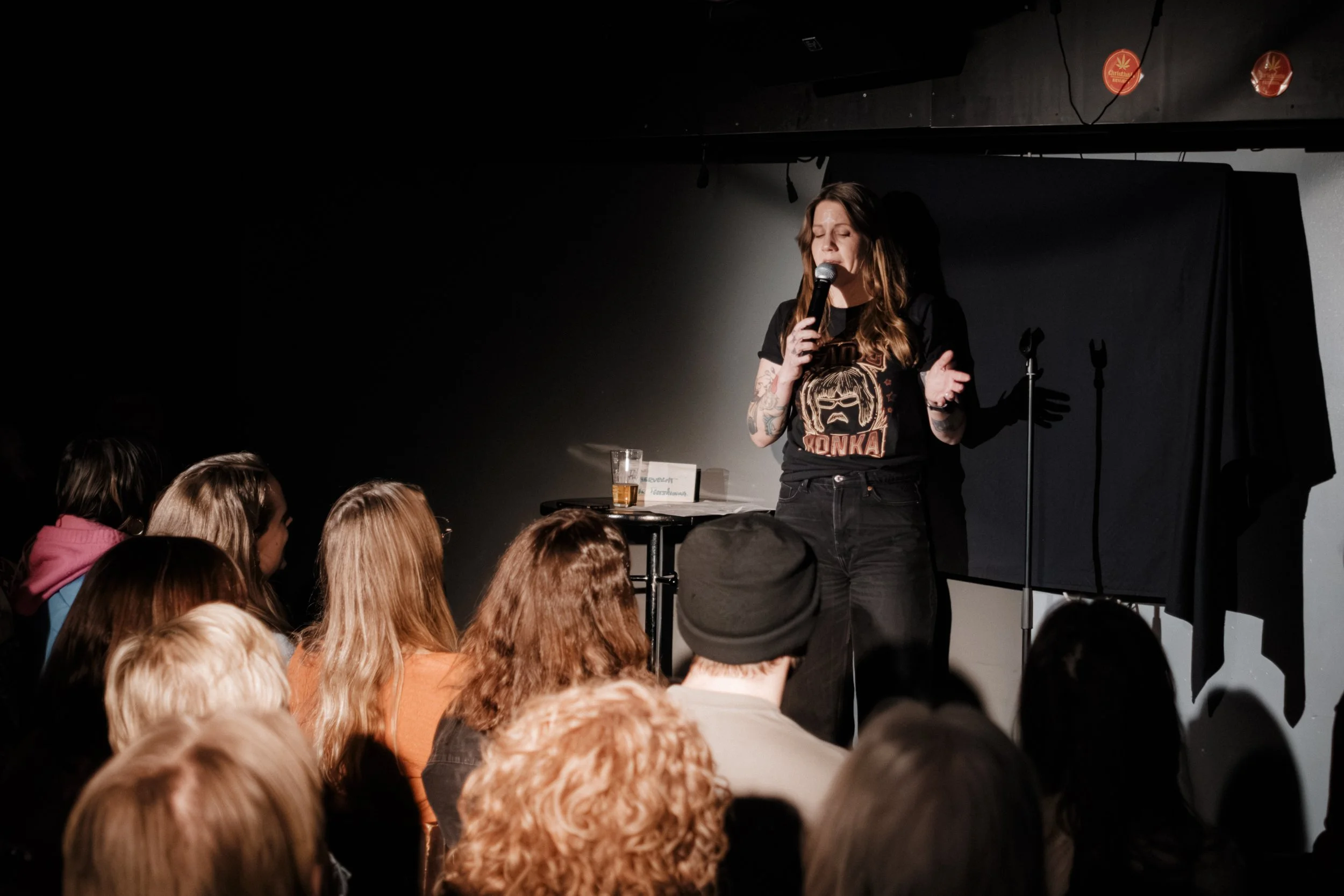 En kvinna som står på en scen och talar till en publik, håller i en mikrofon, med en ölglas framför sig och några mikrofonstativ i bakgrunden.