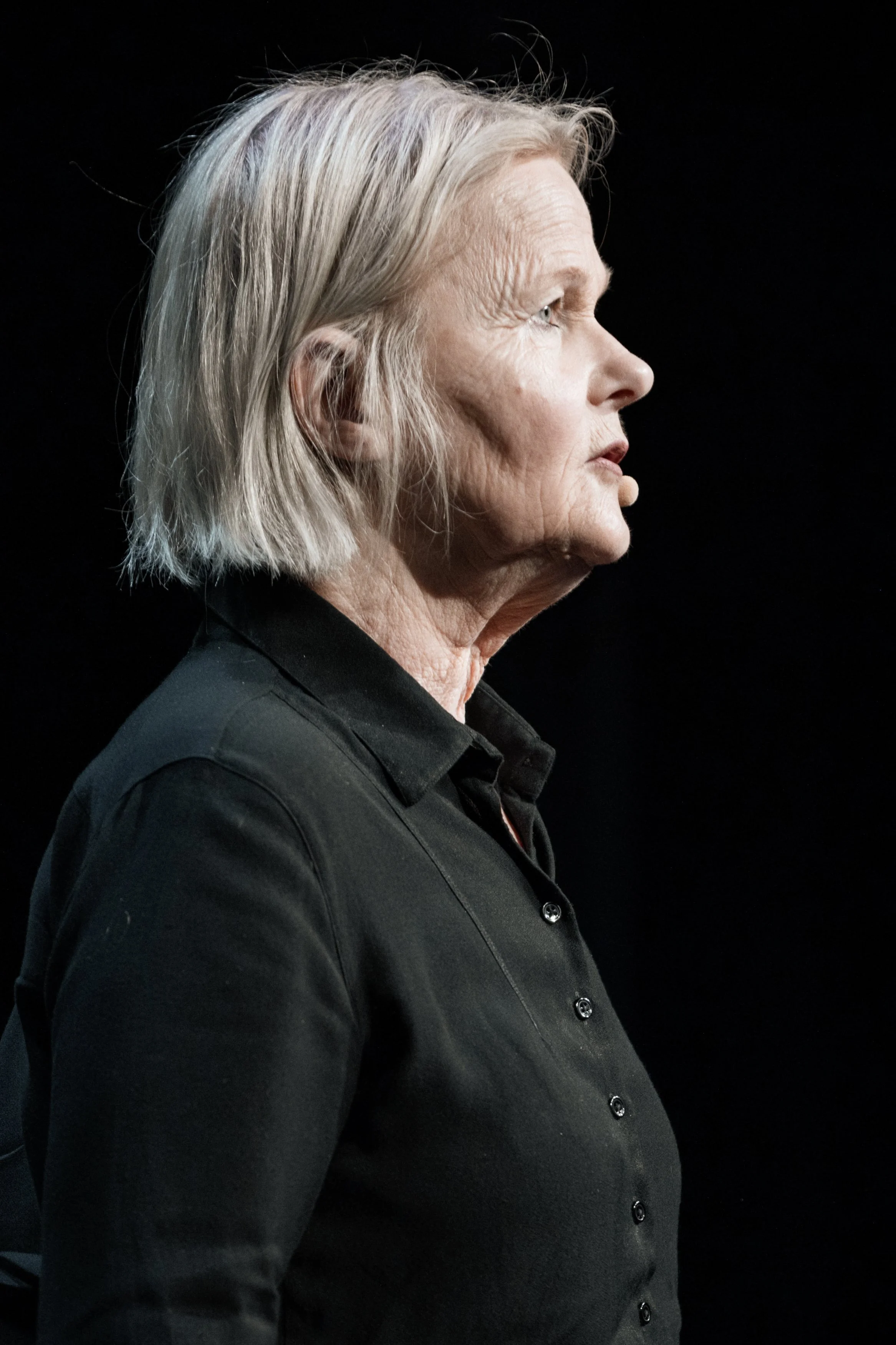 Profilbild av en äldre kvinna med grått hår, klädd i svart skjorta, mot en svart bakgrund.