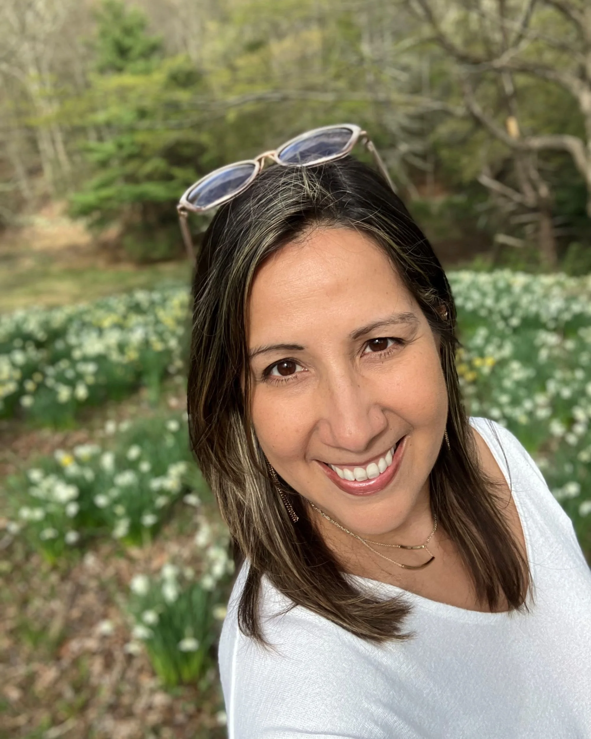 Debra Borrero smiling outdoors in a garden with blooming white flowers, wearing sunglasses on her head and a white top.
