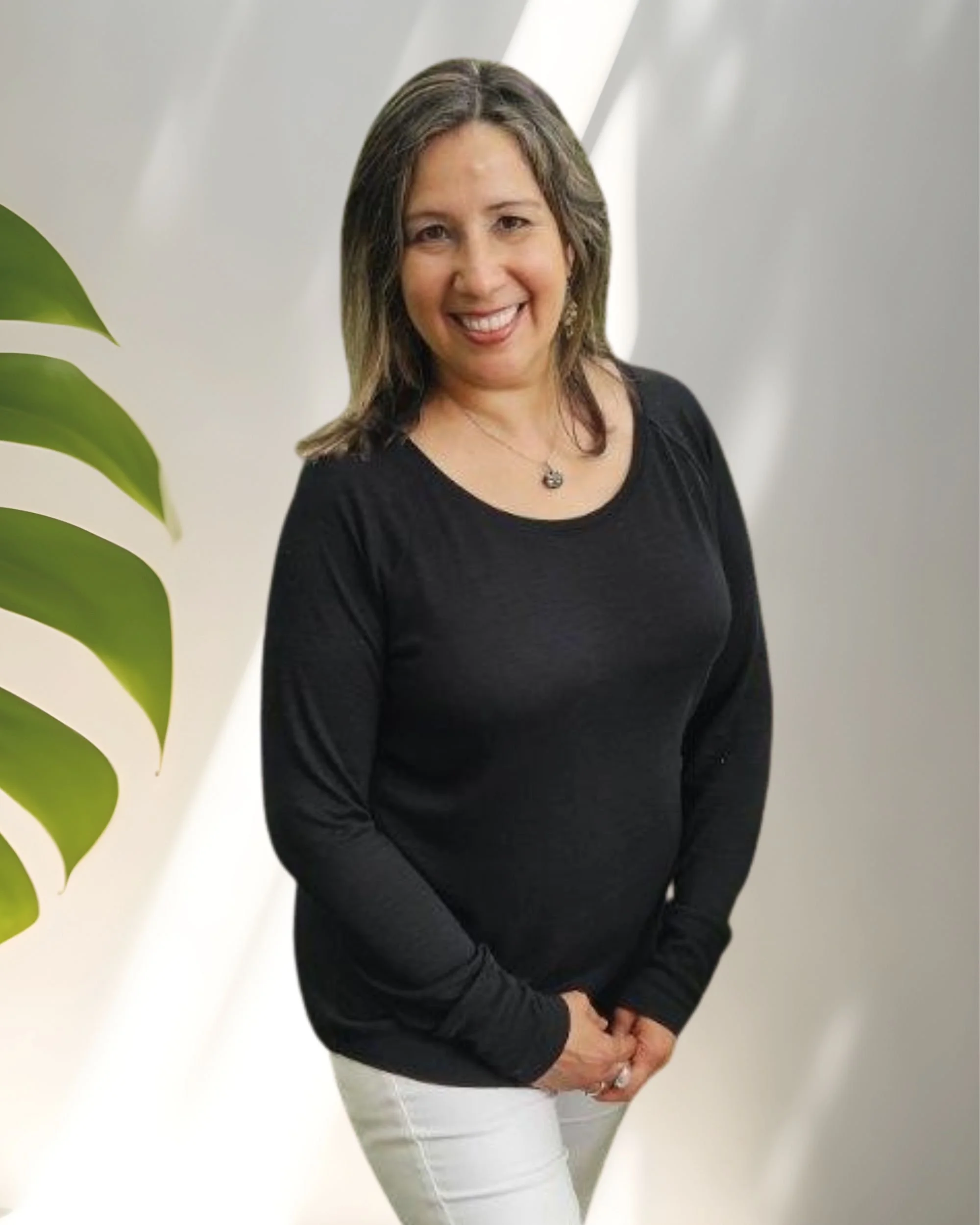 Debra Borrero, woman with shoulder-length brown hair, smiling, wearing a black long-sleeve top and white pants, standing in front of a light-colored background with a green leaf on the left side.