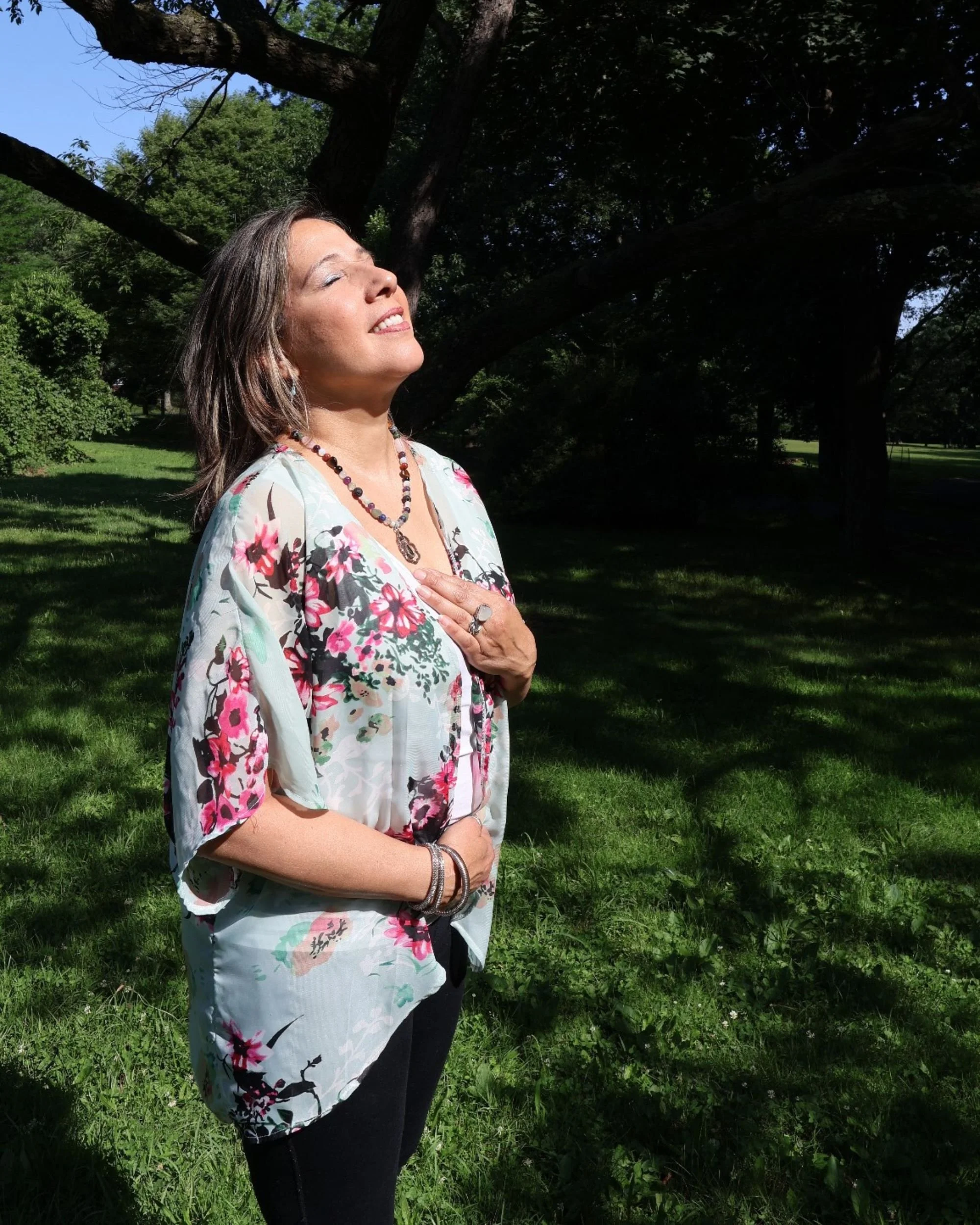 Debra Borrero standing outdoors under a large tree with sunlight shining on her face, eyes closed, wearing a floral blouse and jewelry, with green grass and trees in the background.
