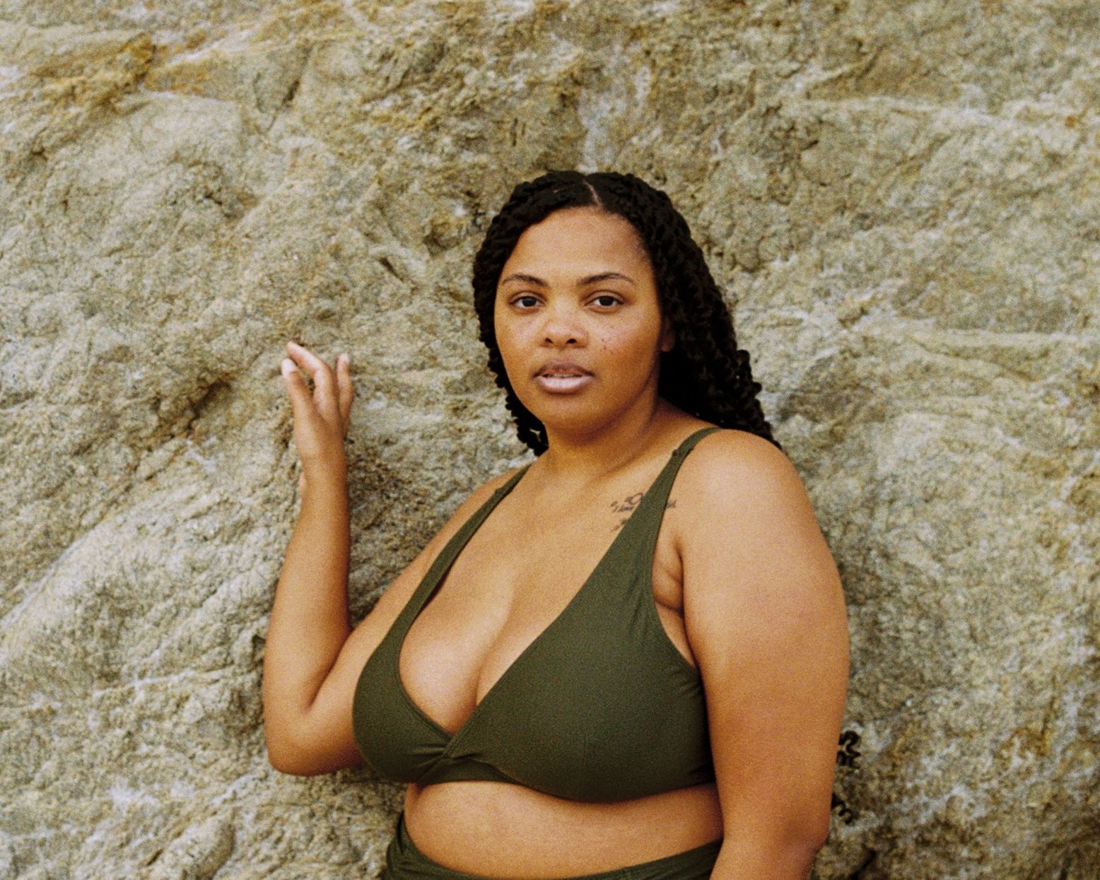 Woman in a green bikini standing against a rock wall with her hand on the rock.