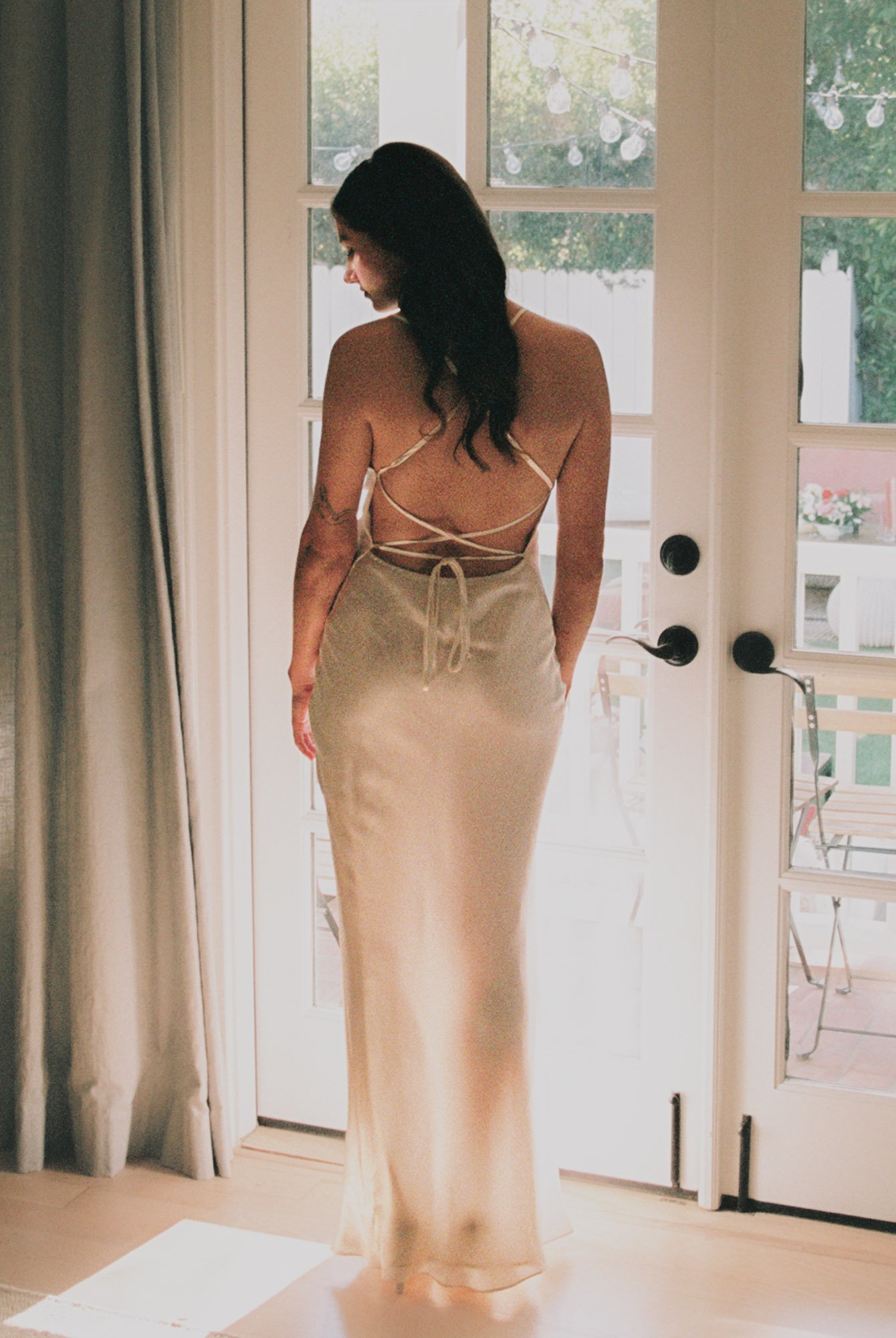 Woman in a beige satin dress with a backless, strappy top standing in front of a glass door, looking downward with natural light.
