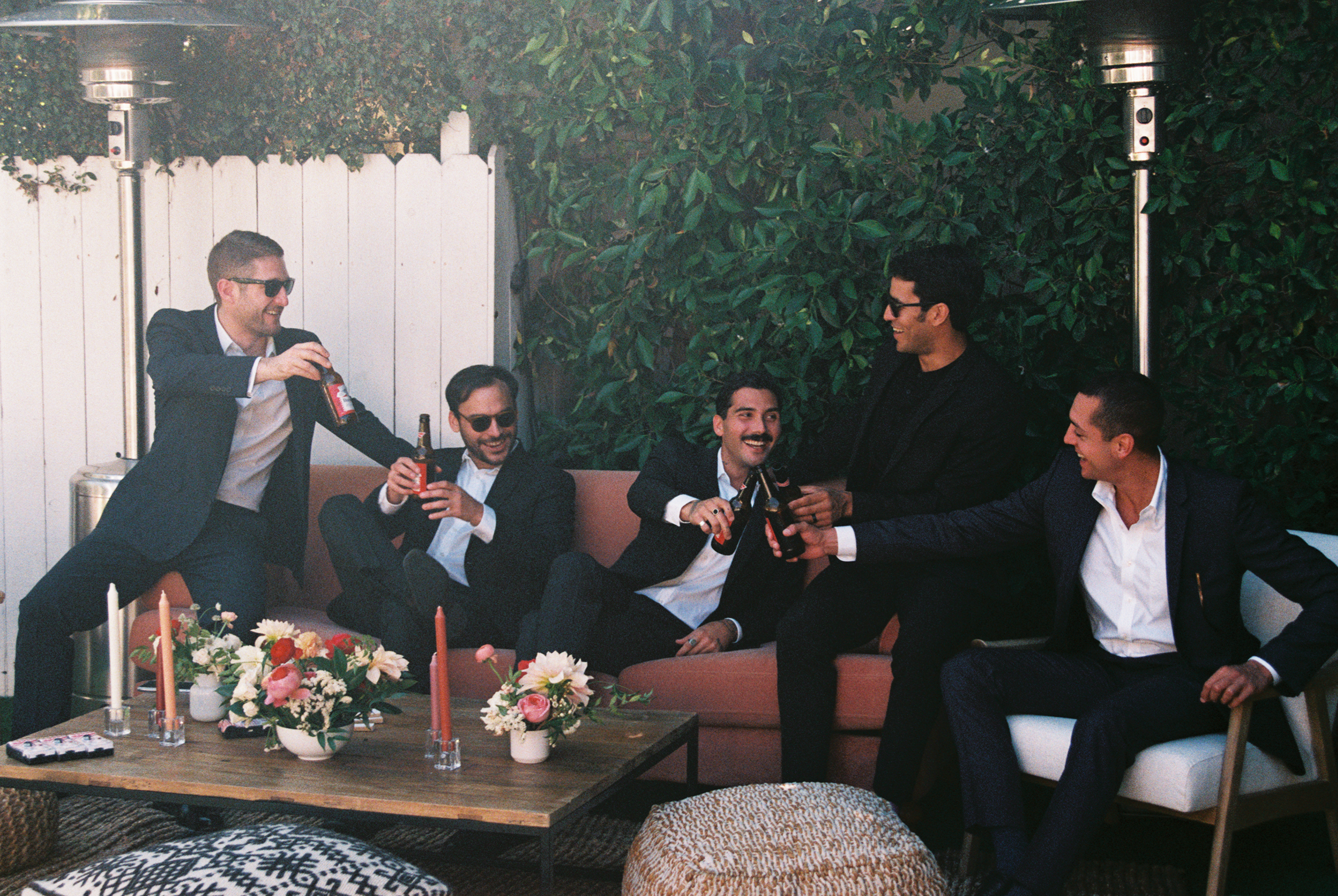 Group of five men wearing suits having fun and drinking beers at an outdoor party, sitting and standing around a wooden table with floral arrangements, candles, and decorative pillows, with a white fence and green foliage in the background.