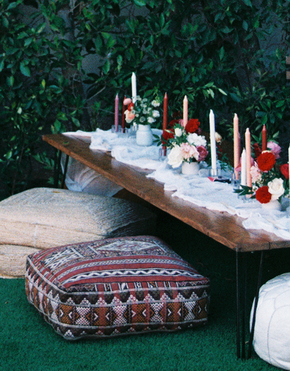 A table decorated with candles and floral arrangements, surrounded by cushions on a grassy surface.