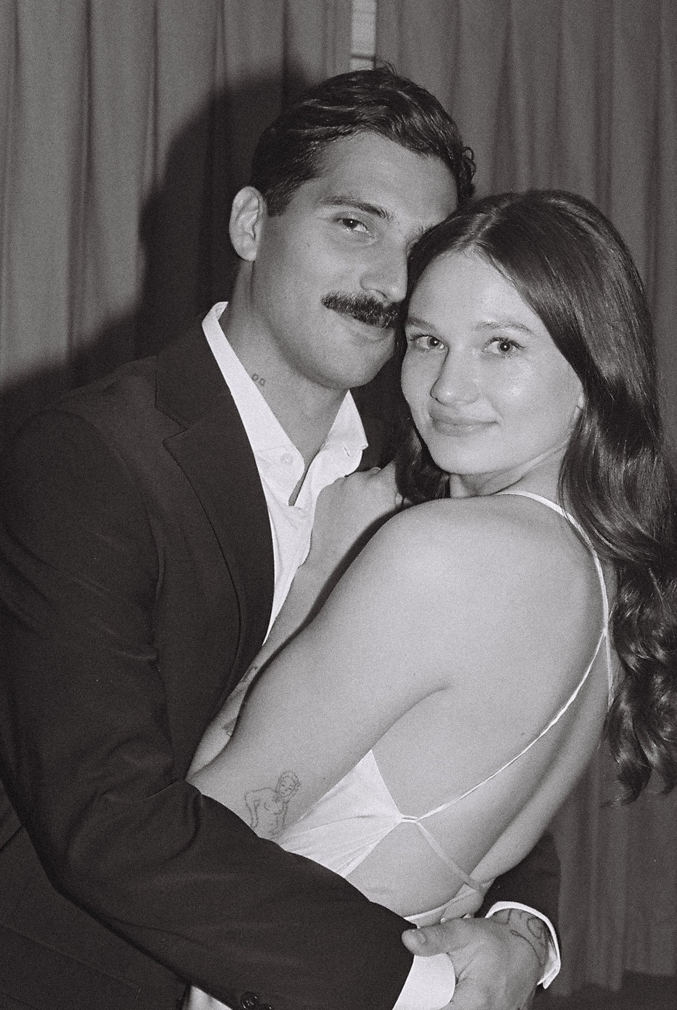 A black and white photo of a smiling couple hugging. The man has a mustache, is wearing a suit, and has dark hair. The woman has long wavy hair, is wearing a sleeveless dress, and has a tattoo on her upper arm.