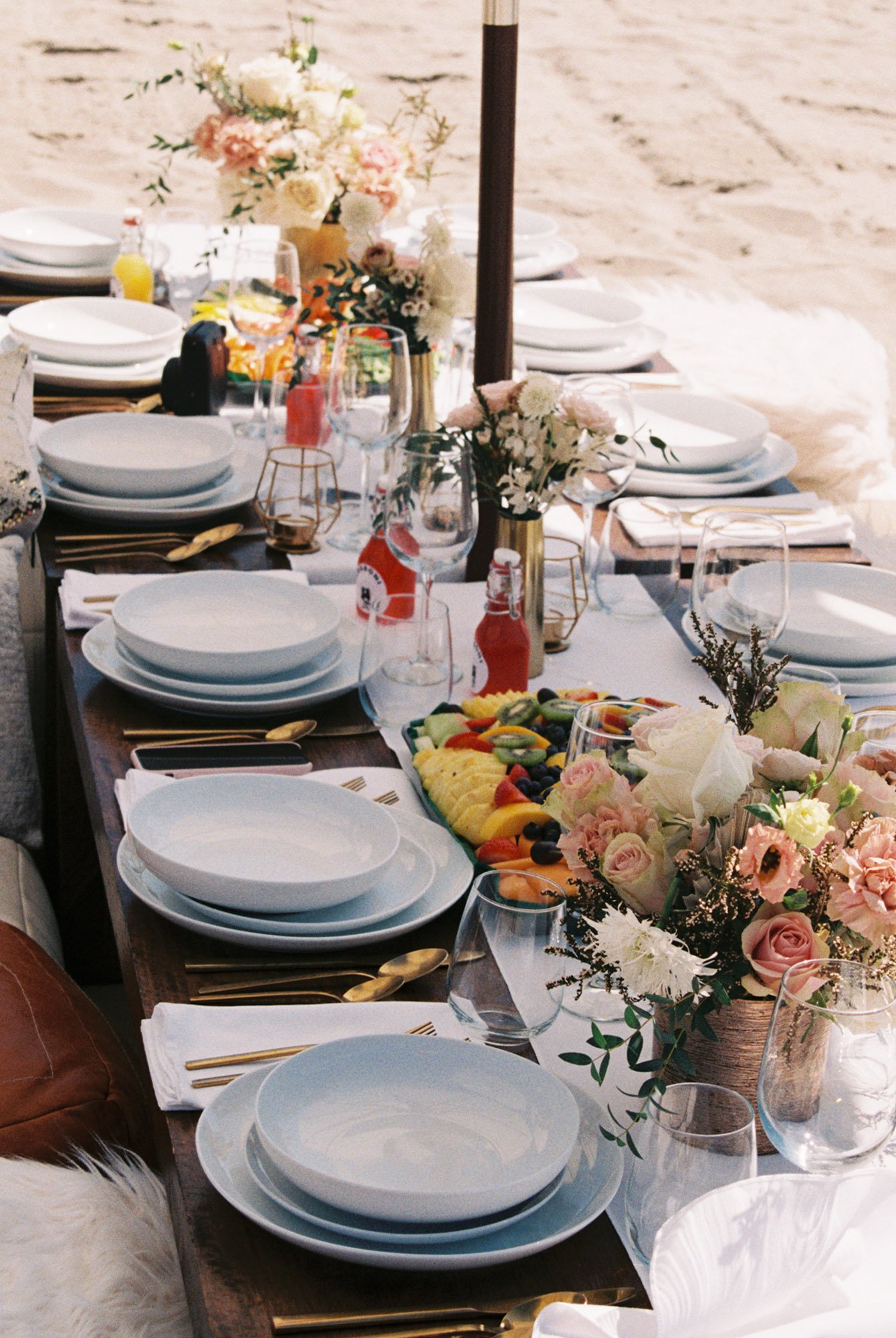 Beachside outdoor dining table with white plates, gold cutlery, wine glasses, floral centerpieces, and a platter of sliced fruit.
