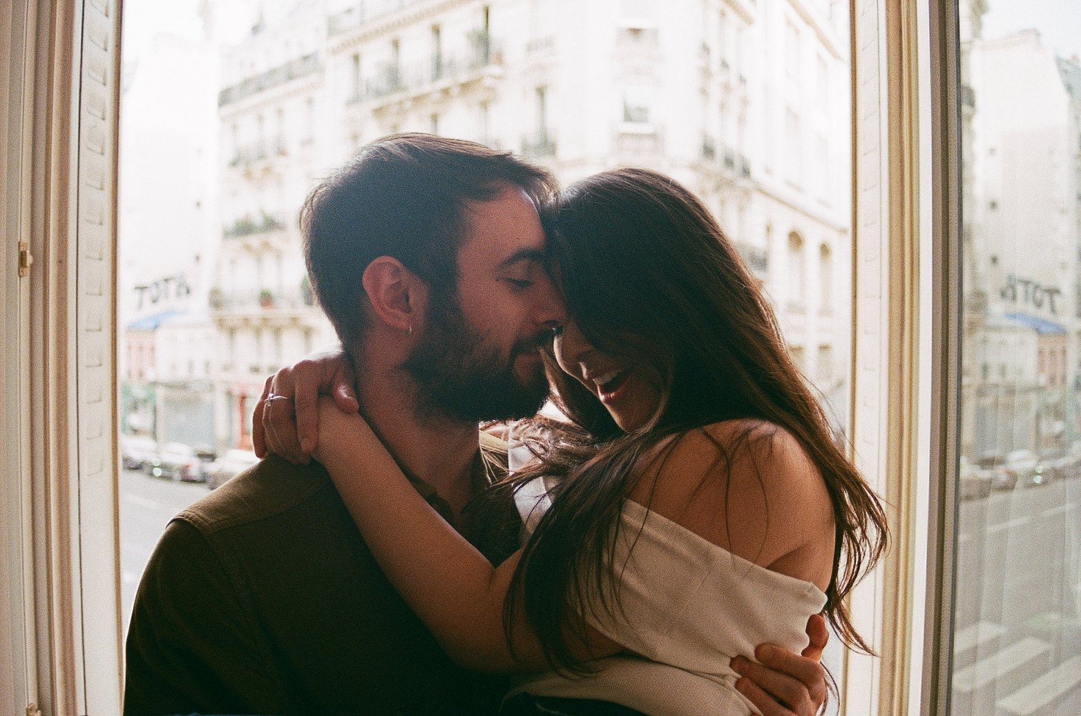 A couple sharing a joyful embrace and laughter by a large window with a cityscape background.