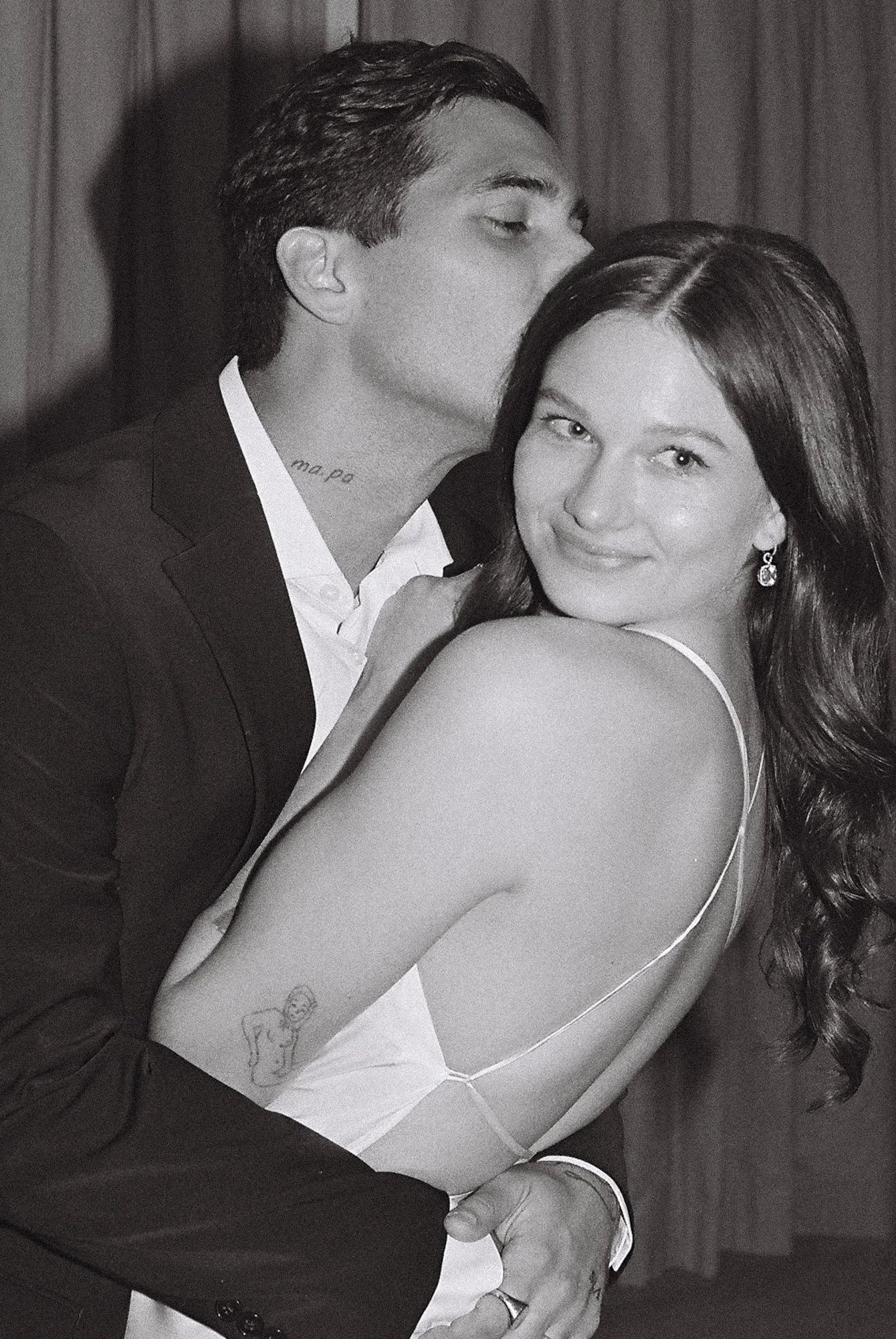 A man in a suit kisses a woman on the temple while holding her from behind. The woman has long dark hair, is smiling at the camera, and is wearing a spaghetti strap dress. The photo is in black and white.