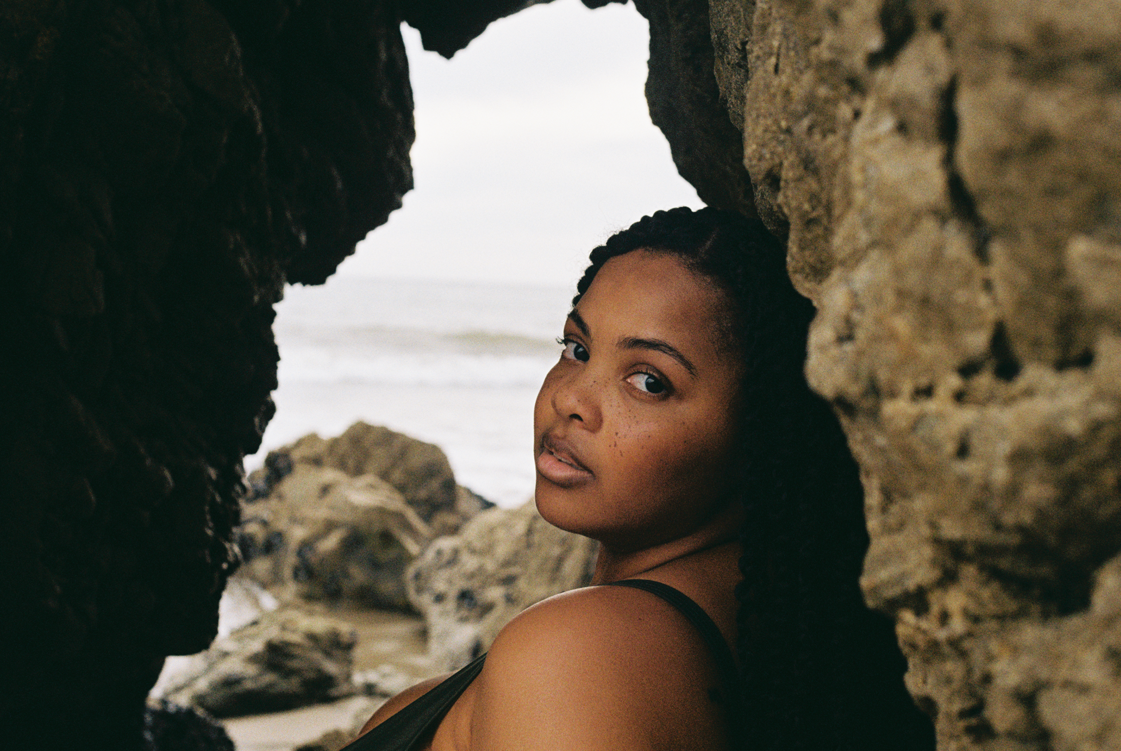 A woman with dark hair and freckles leans against a rocky formation on a beach, looking at the camera with a serious expression.