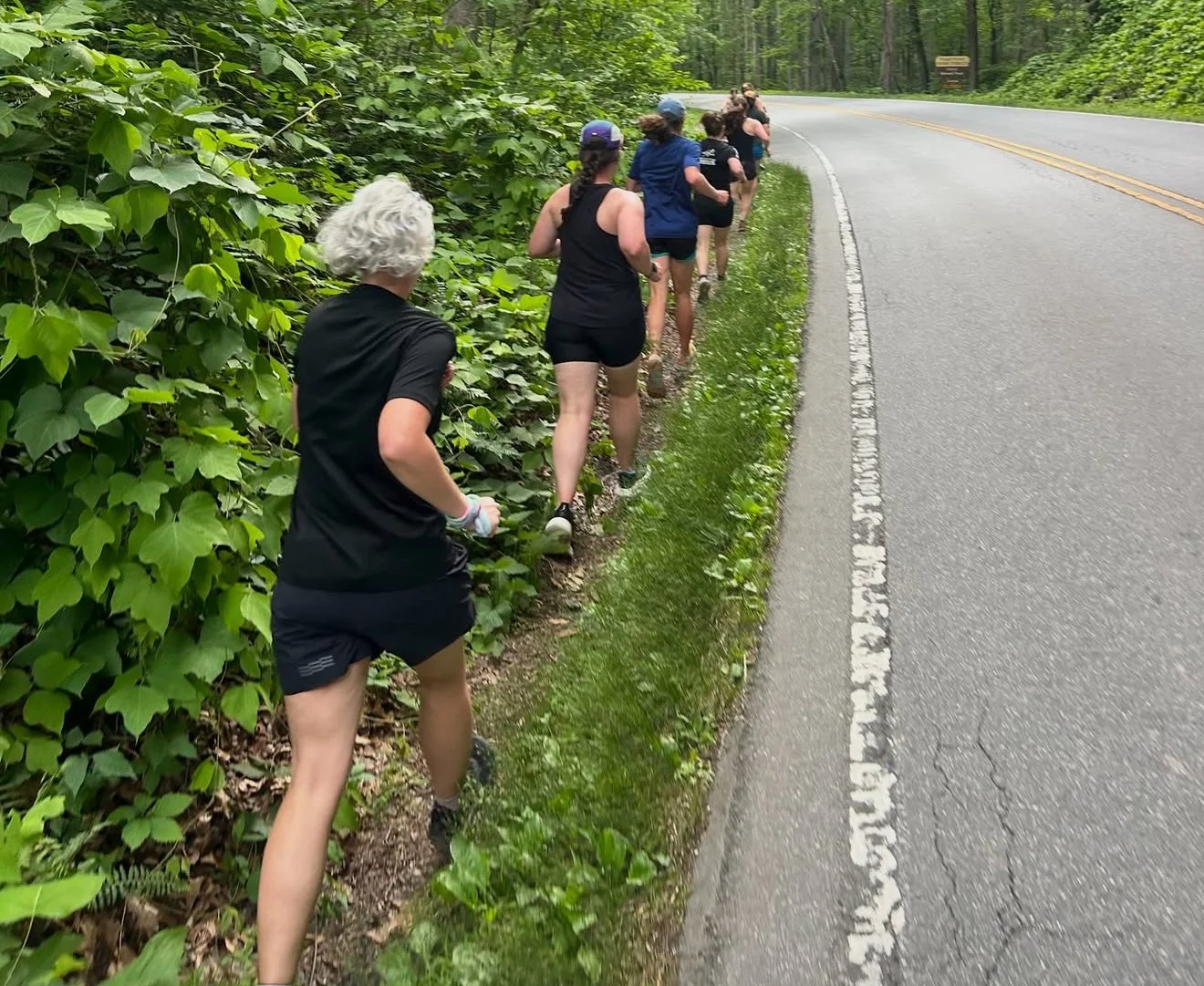 A group of people running on a narrow dirt trail alongside a paved road with forested greenery on one side.