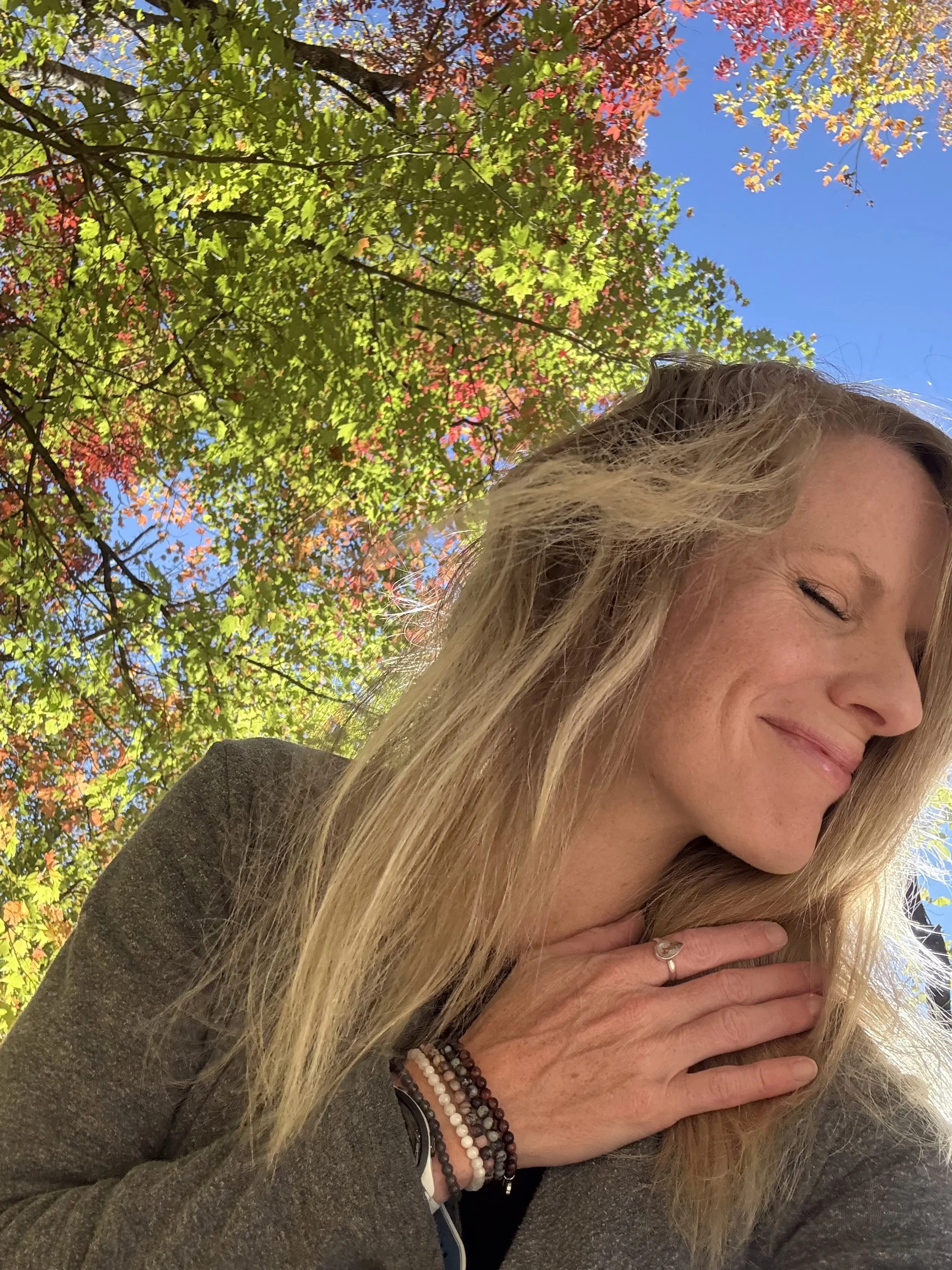 A woman with long blonde hair smiling with her eyes closed, holding her hand to her neck, outdoors on a sunny day with vibrant fall foliage in green, red, and orange colors against a bright blue sky.