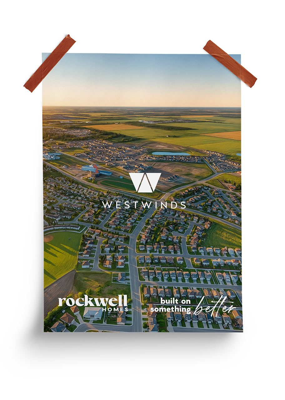 Elevated aerial view of a residential neighborhood with houses, green fields, a pond, and open fields in the background, under a clear sky during sunset. The image features the Westwinds logo and slogan 'Rockwell Homes built on something better'.