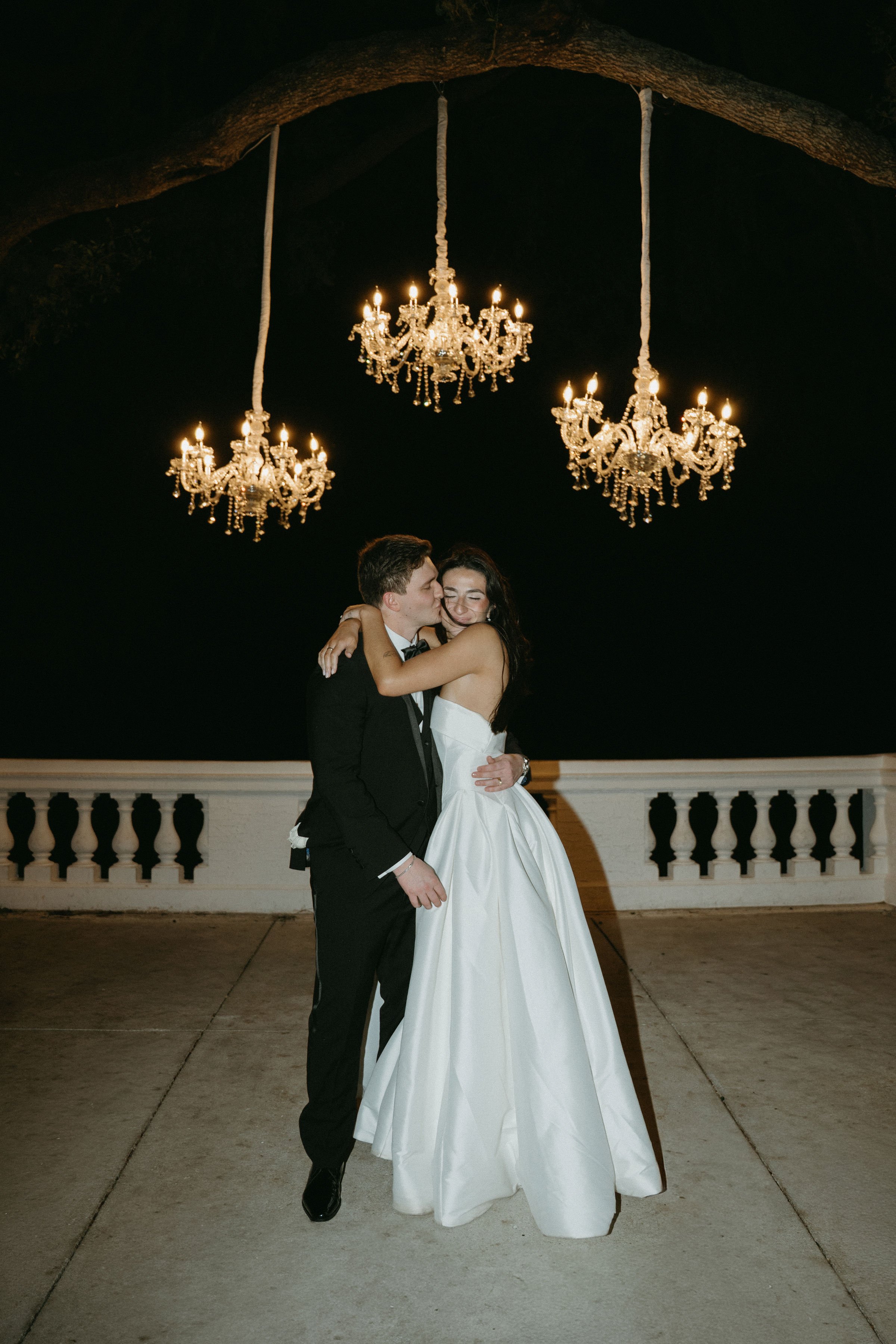 Bella and Juan embracing during their private last dance beneath three glowing chandeliers outside Bella Cosa at night.