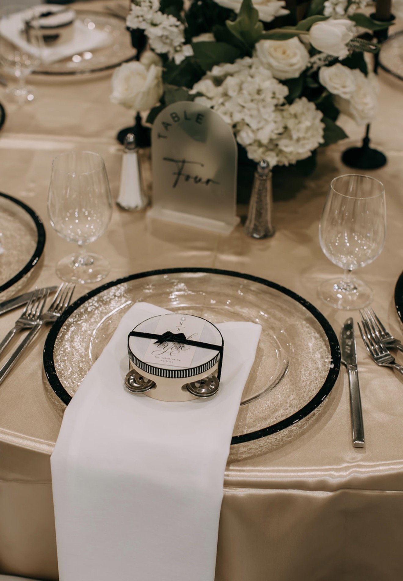 Place setting with clear charger plate with black rim, champagne satin tablecloth, white cloth napkin, thank you card, and tambourine wedding favor.