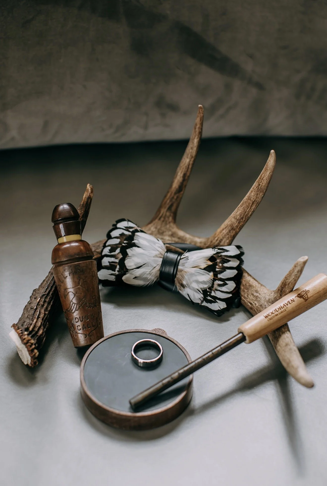 Groom’s detail flat lay with pheasant feather bow tie, antler shed, turkey call, and duck call representing his love of hunting.