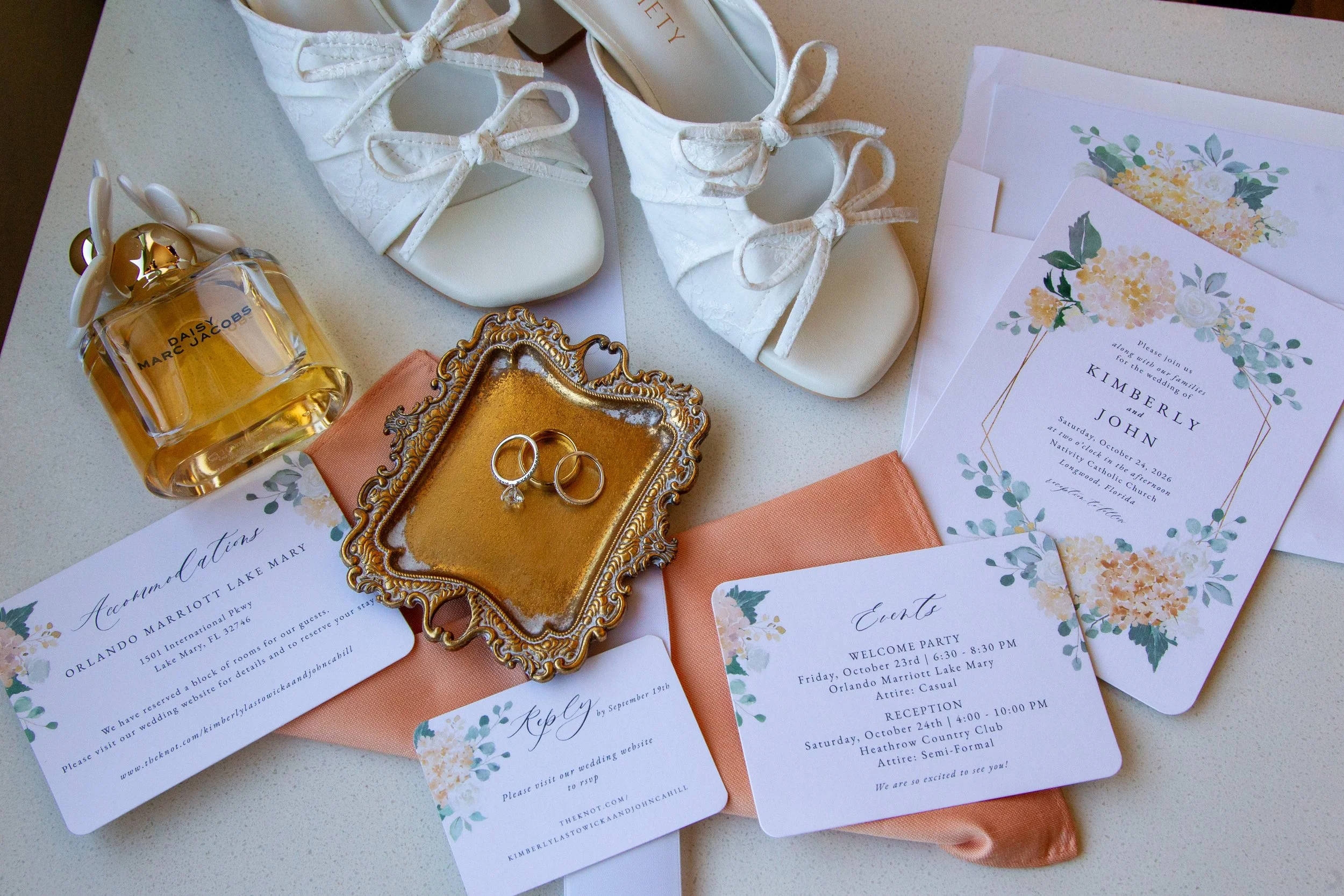 Floral wedding invitations, white bridal shoes, gold wedding rings, Daisy perfume, and peach fabric arranged in a wedding detail flat lay.