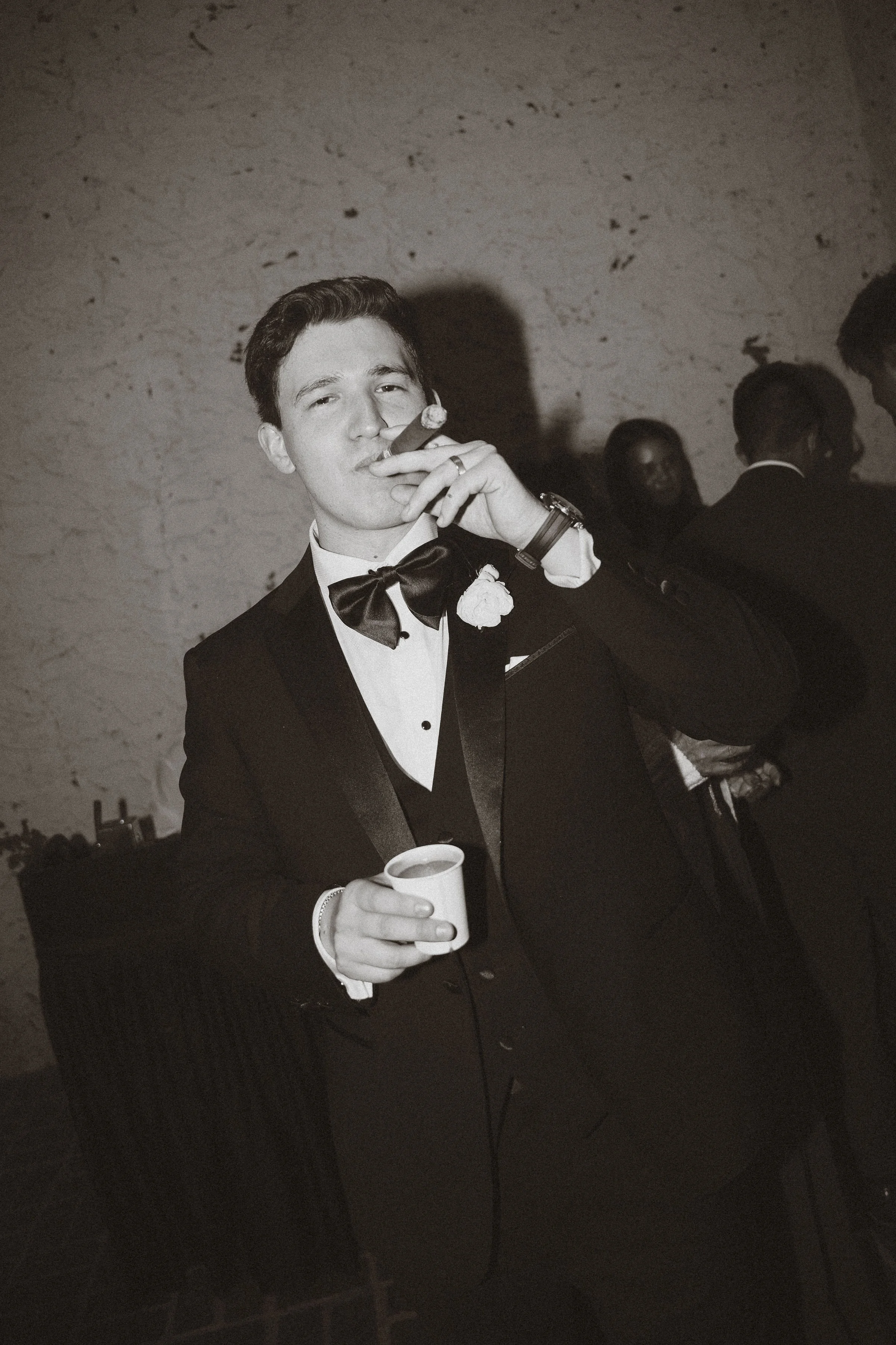 Juan wearing a black tuxedo and holding a cigar and small cup of Cuban coffee during his wedding reception.