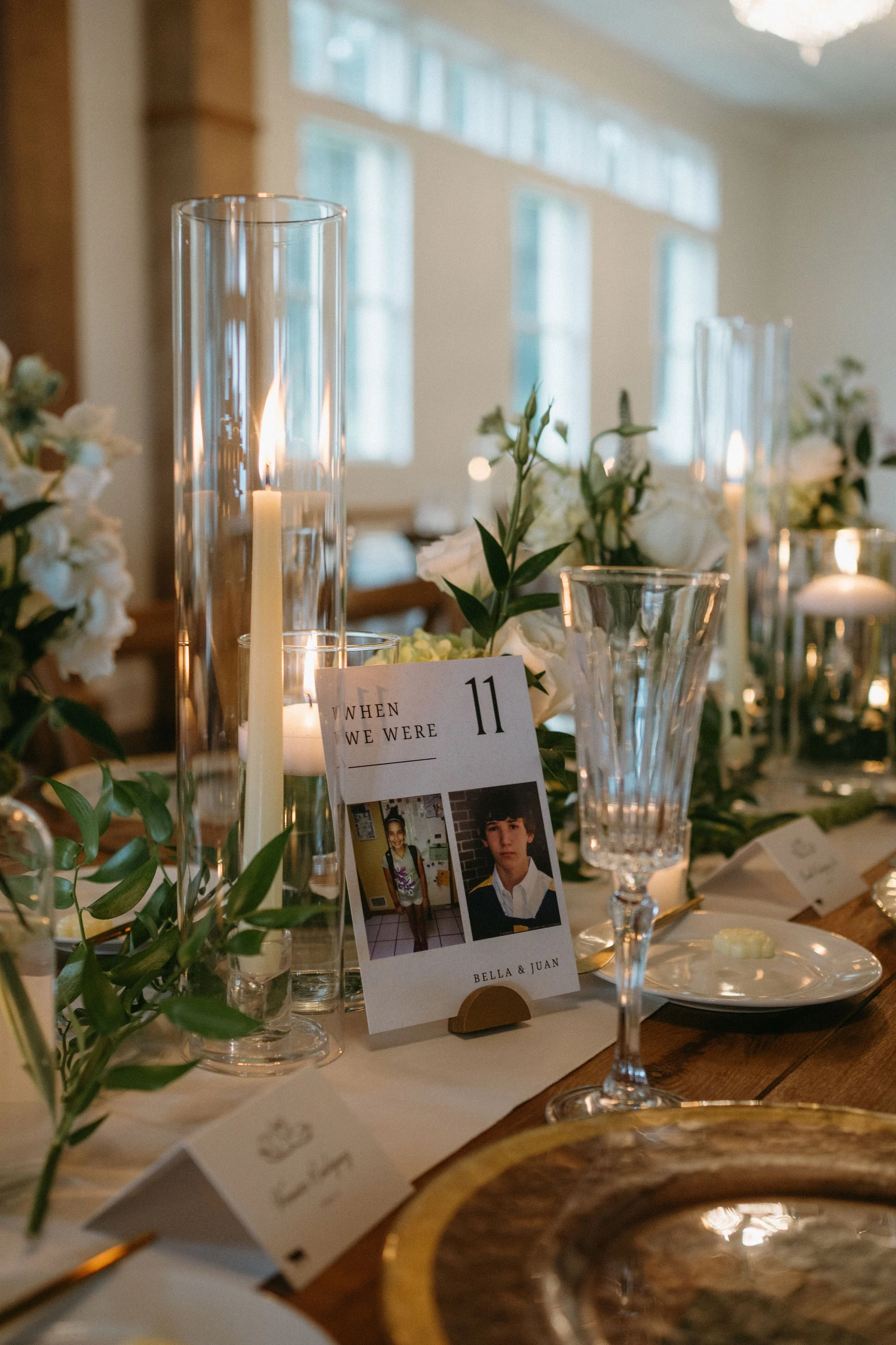 Wedding reception table with candles, white flowers, greenery, gold charger, and table number eleven featuring childhood photos of Bella and Juan.