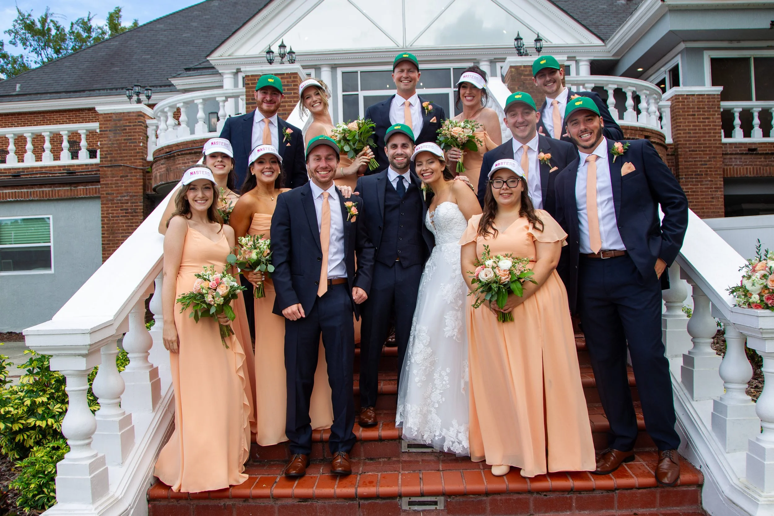 Kimberly and John posing with their wedding party in Masters golf hats, peach bridesmaid dresses, and navy suits on the steps of Heathrow Country Club.