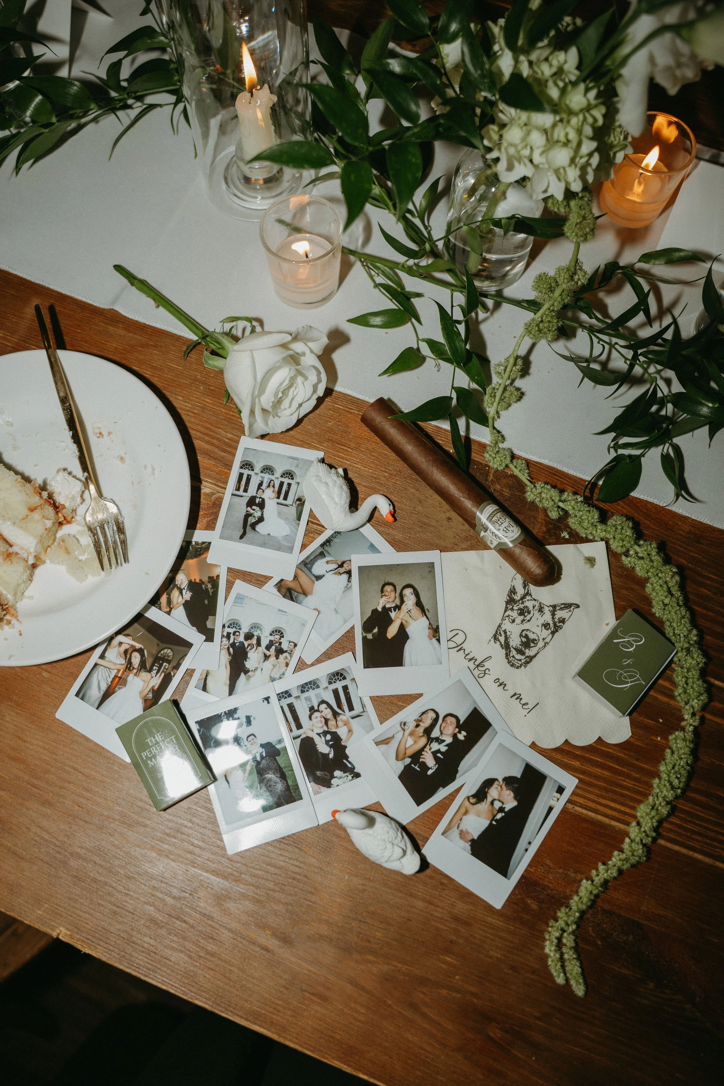 Wedding detail photograph with Polaroid guest pictures, white swan figurines, a cigar, white roses, and candlelight.