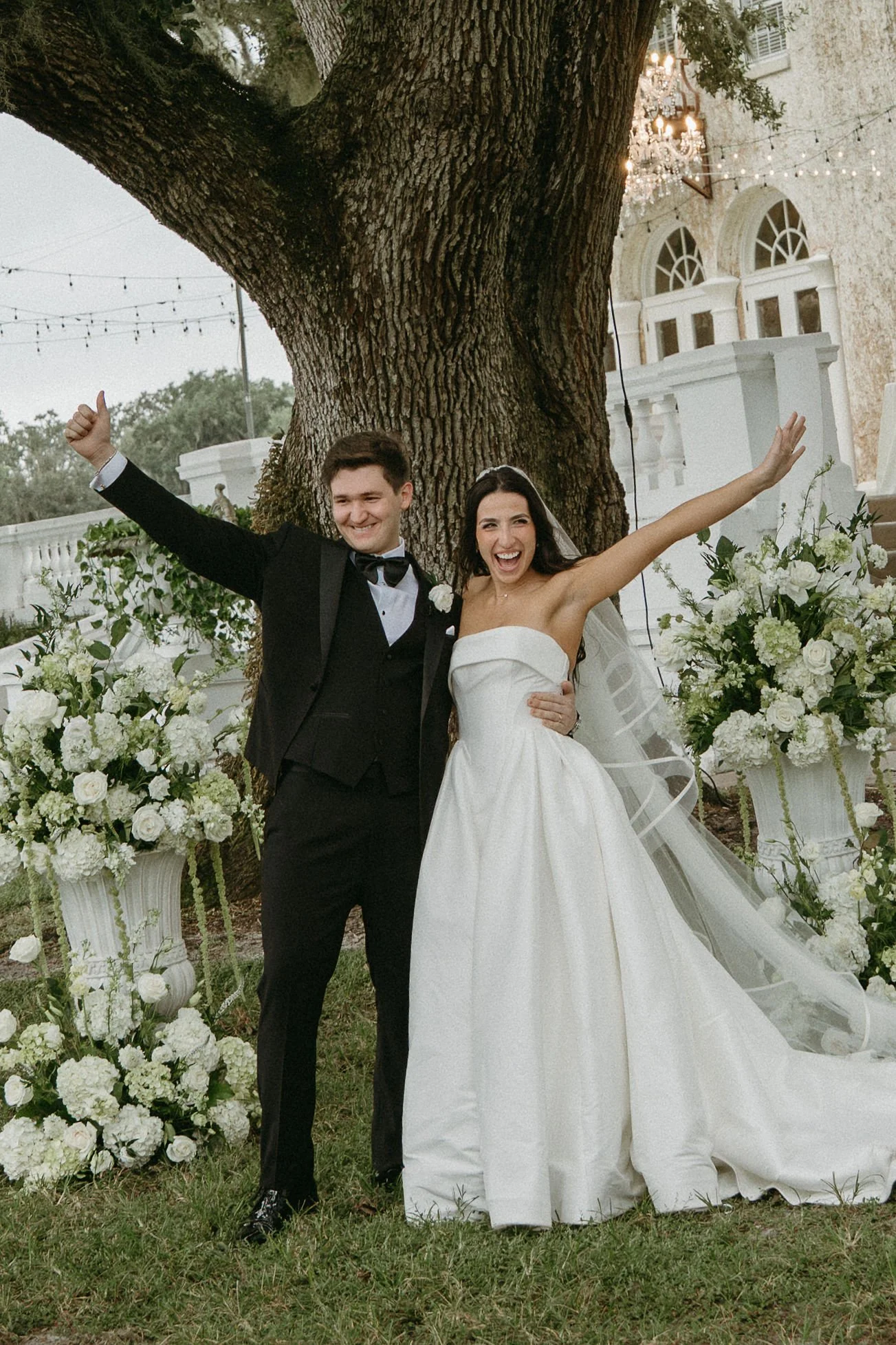 Bella and Juan smiling with their arms raised in celebration beneath the oak tree after their outdoor wedding ceremony.