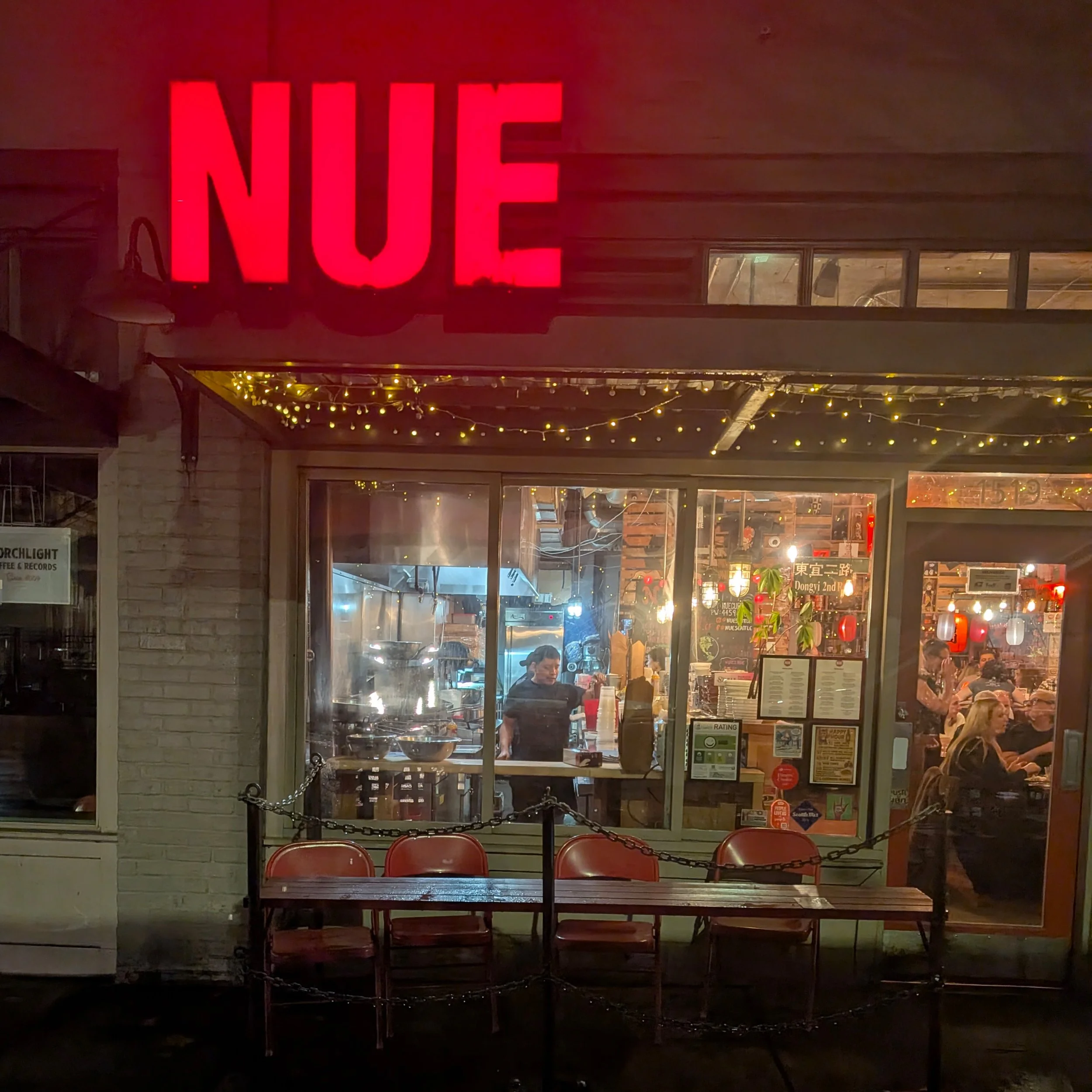 Night view of a storefront with a large bright red neon sign that says 'NUE', with interior lighting visible through the windows, and decorated string lights hanging outside. There are chairs and people inside the restaurant.